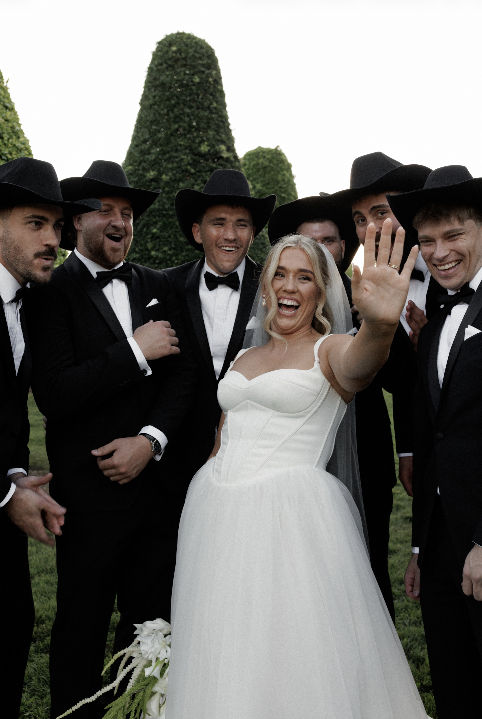Bride and groom with friends in tuxedos and cowboy hats, outdoors during daytime.