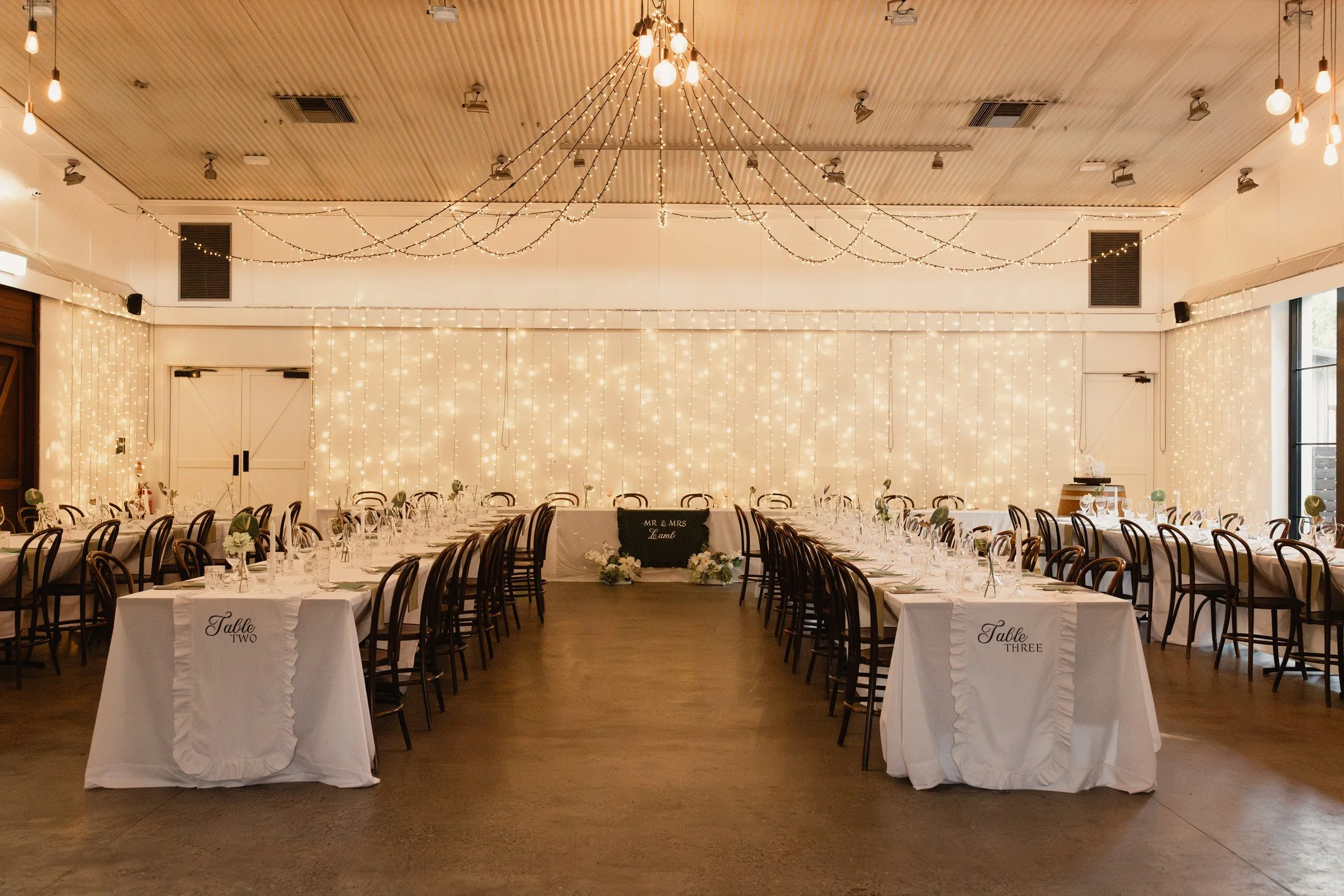 A banquet hall decorated with string lights and curtains for a wedding reception, with tables set for guests.