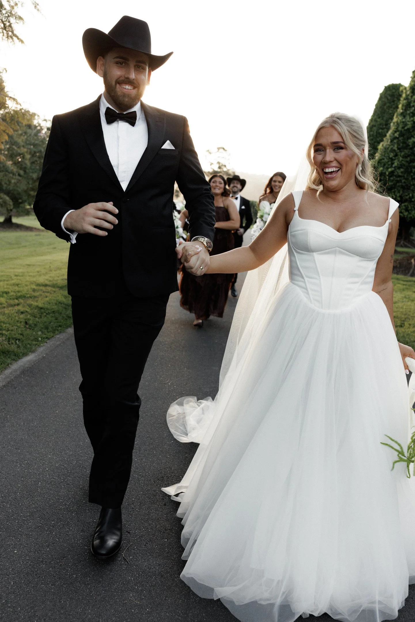 A groom and bride celebrating their wedding outdoors, holding hands and smiling. The groom is dressed in a black tuxedo with a bow tie and cowboy hat, while the bride is in a white wedding gown with a veil. Other wedding guests are visible in the bac