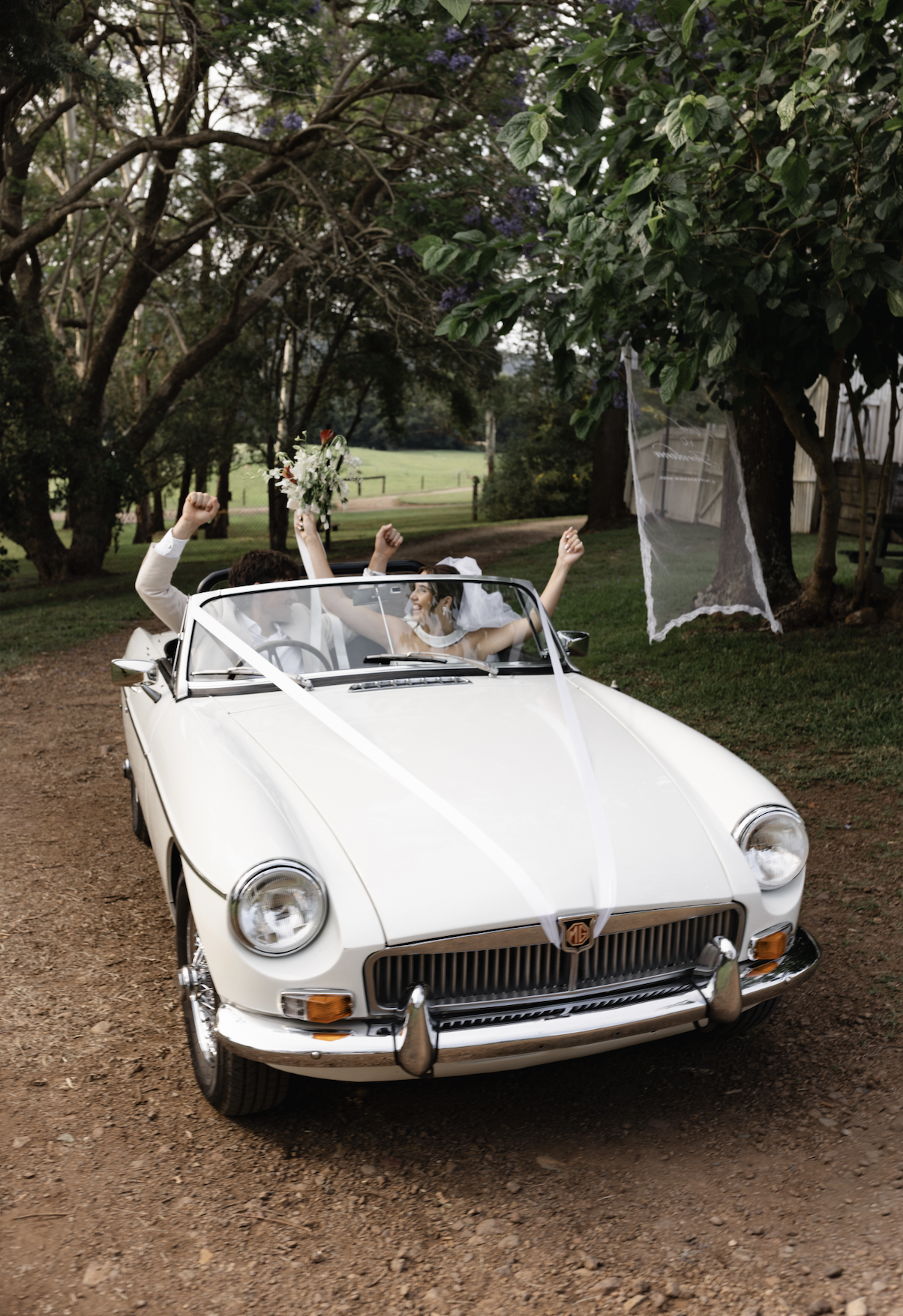 A newlywed couple celebrating in a vintage white MG convertible car outdoors surrounded by trees, with the bride holding a bouquet and both raising their fists in joy.