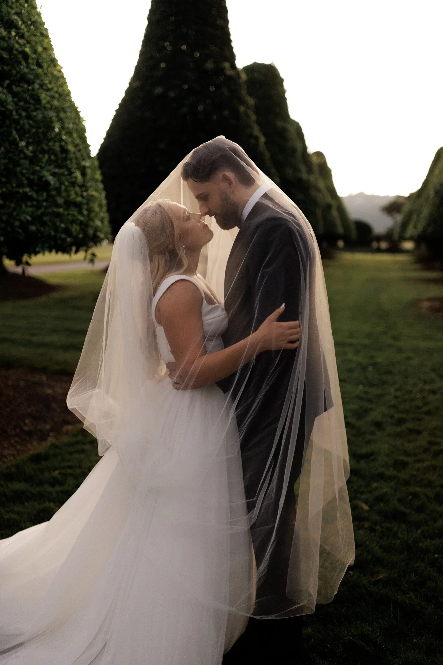 A bride and groom stand closely together outdoors, dressed in wedding attire, with the bride wearing a white gown and veil, and the groom in a dark suit, gazing into each other's eyes under a veil, surrounded by tall, lush green trees.