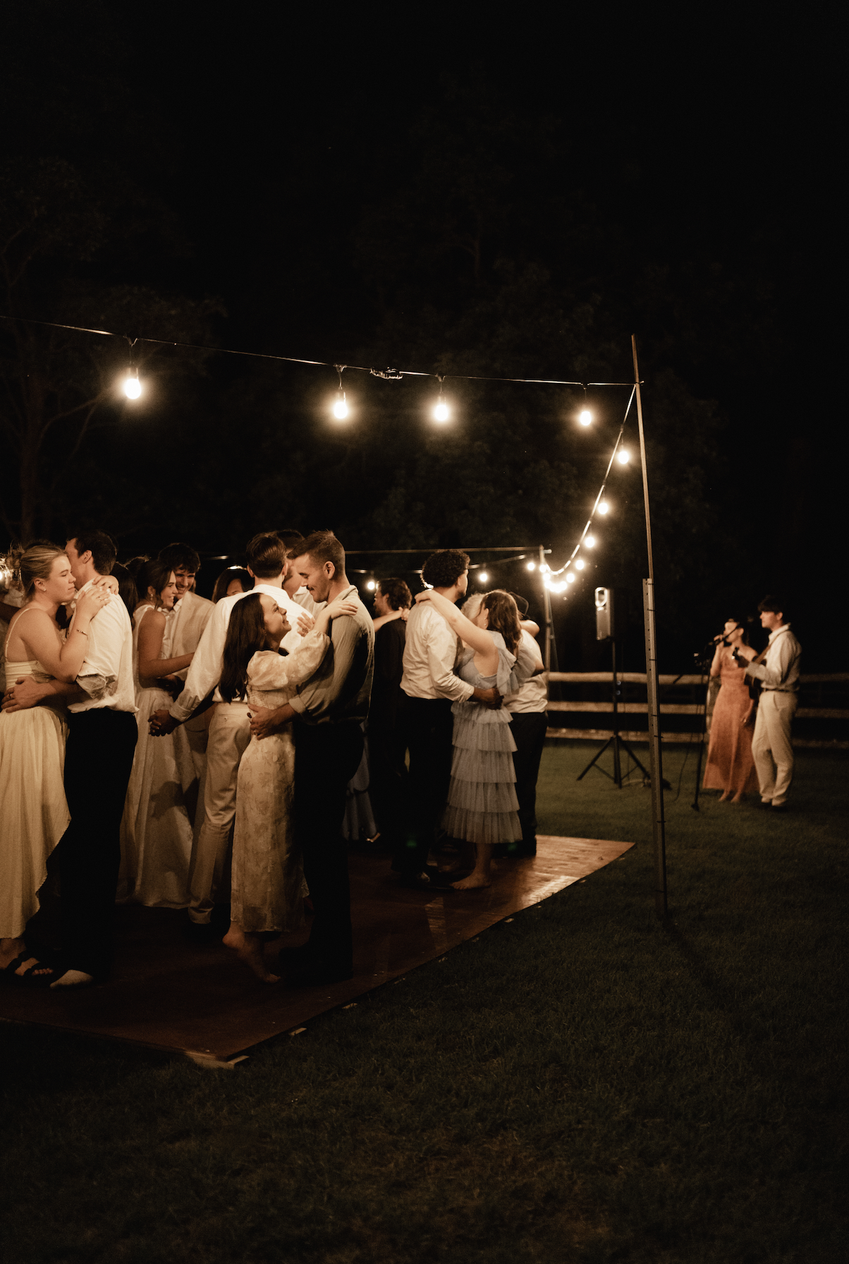 A group of people dancing and socializing on an outdoor wooden dance floor at night, under hanging string lights, with a couple singing at a microphone in the background.