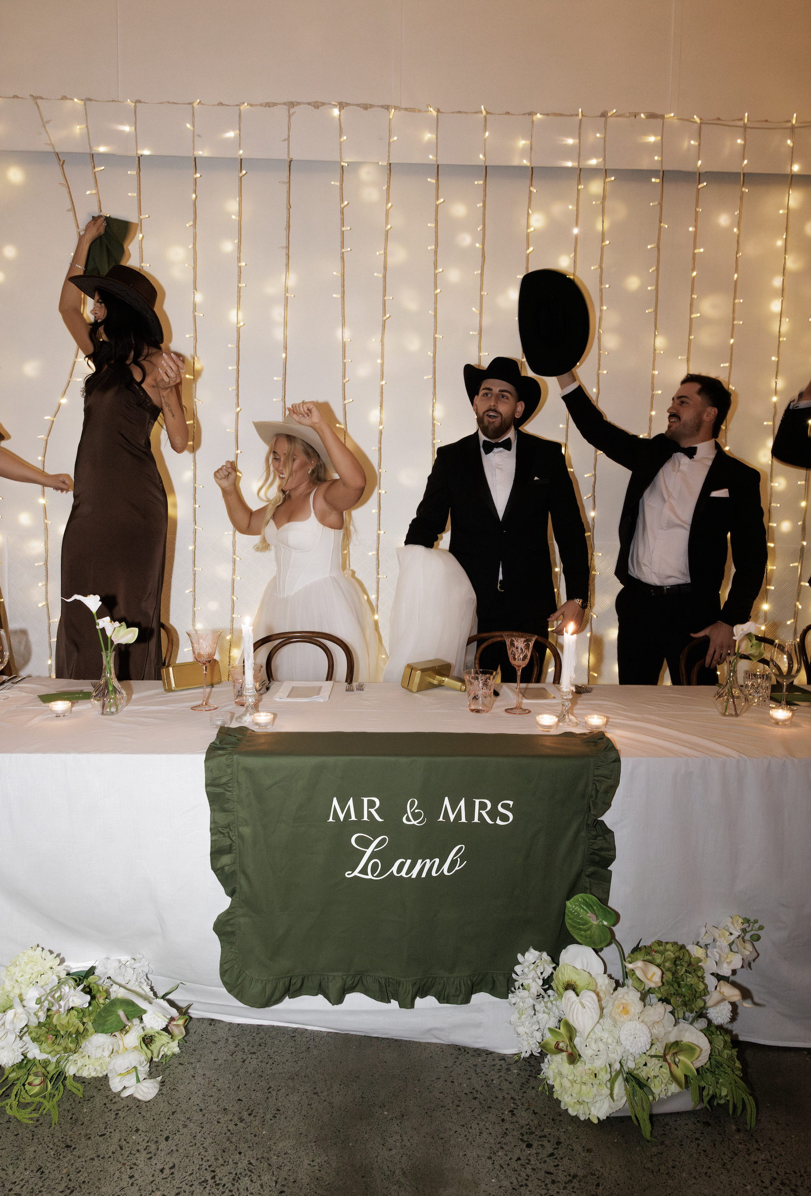 People at a wedding reception, with a head table decorated with flowers and a green and white banner reading "Mr & Mrs Lamb." The backdrop features string lights, and some guests are wearing hats and dancing.