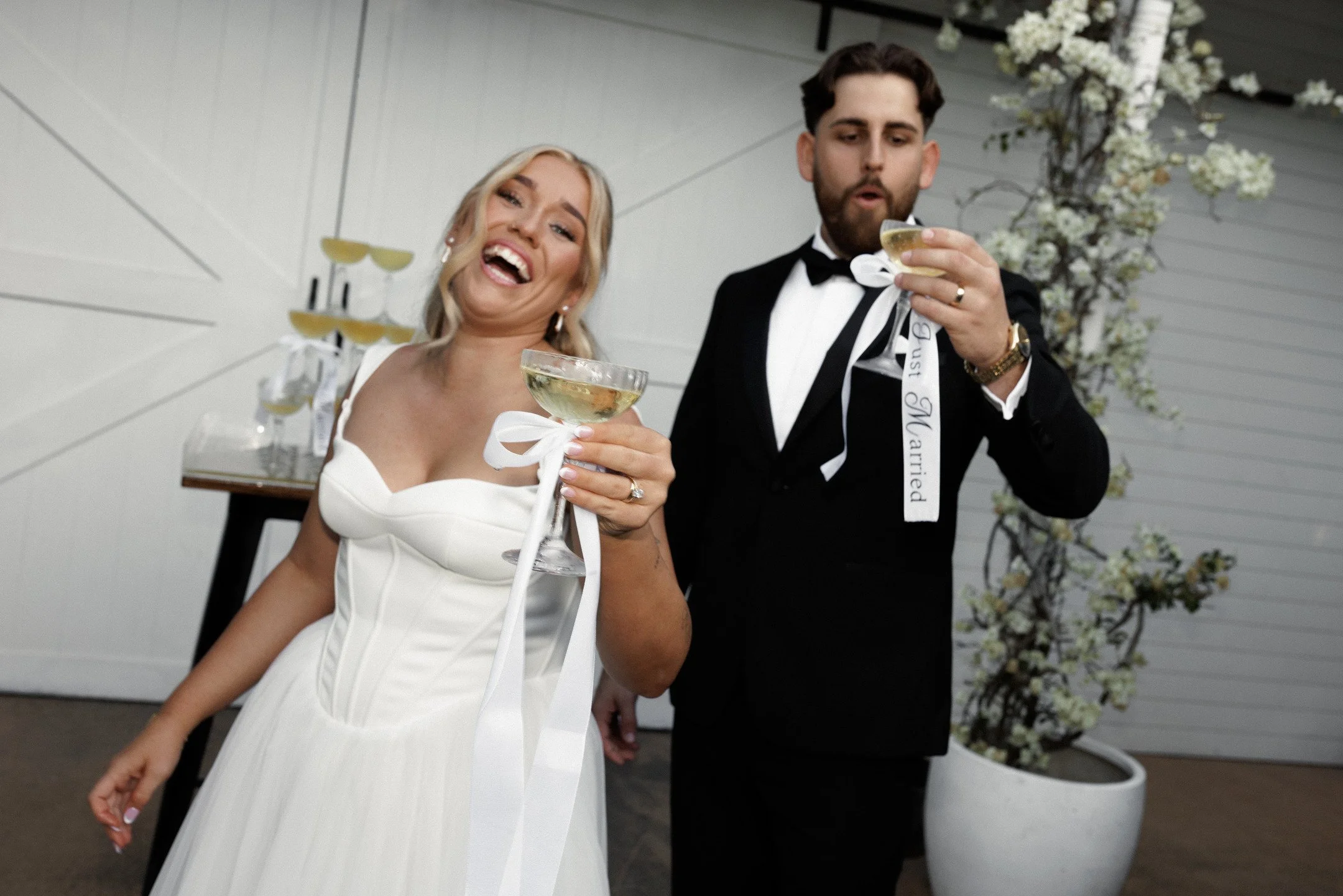 A newlywed couple at their wedding reception, raising glasses in a toast, smiling and enjoying the celebration indoors with floral decorations in the background.