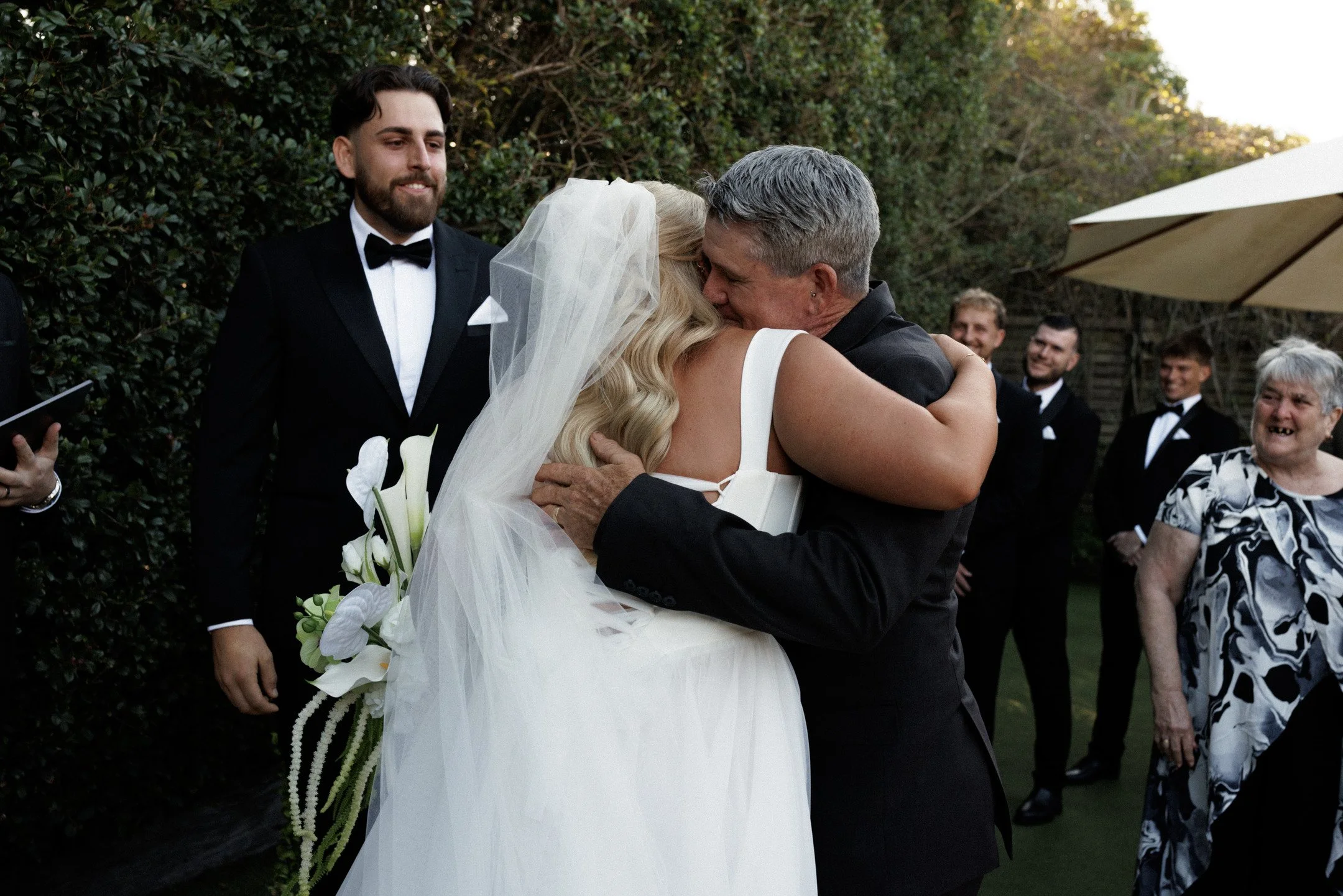 A bride and an older man, possibly her father, hugging at an outdoor wedding ceremony. The bride is holding white calla lilies and wearing a wedding gown and veil. Guests in tuxedos and formal attire are smiling in the background.