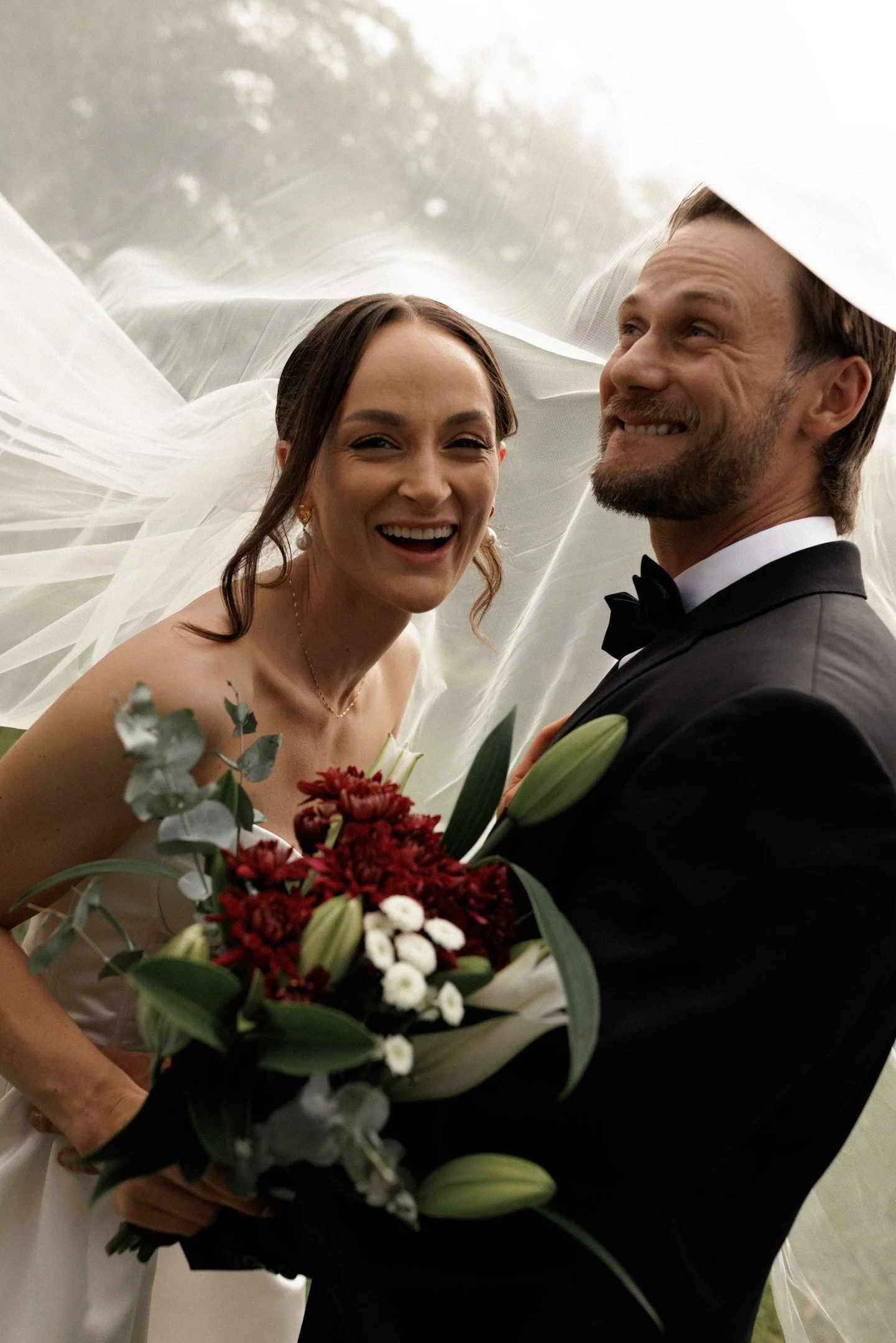 A smiling bride and groom on their wedding day, with the bride holding a bouquet of red and white flowers, and the veil flowing in the wind behind them.
