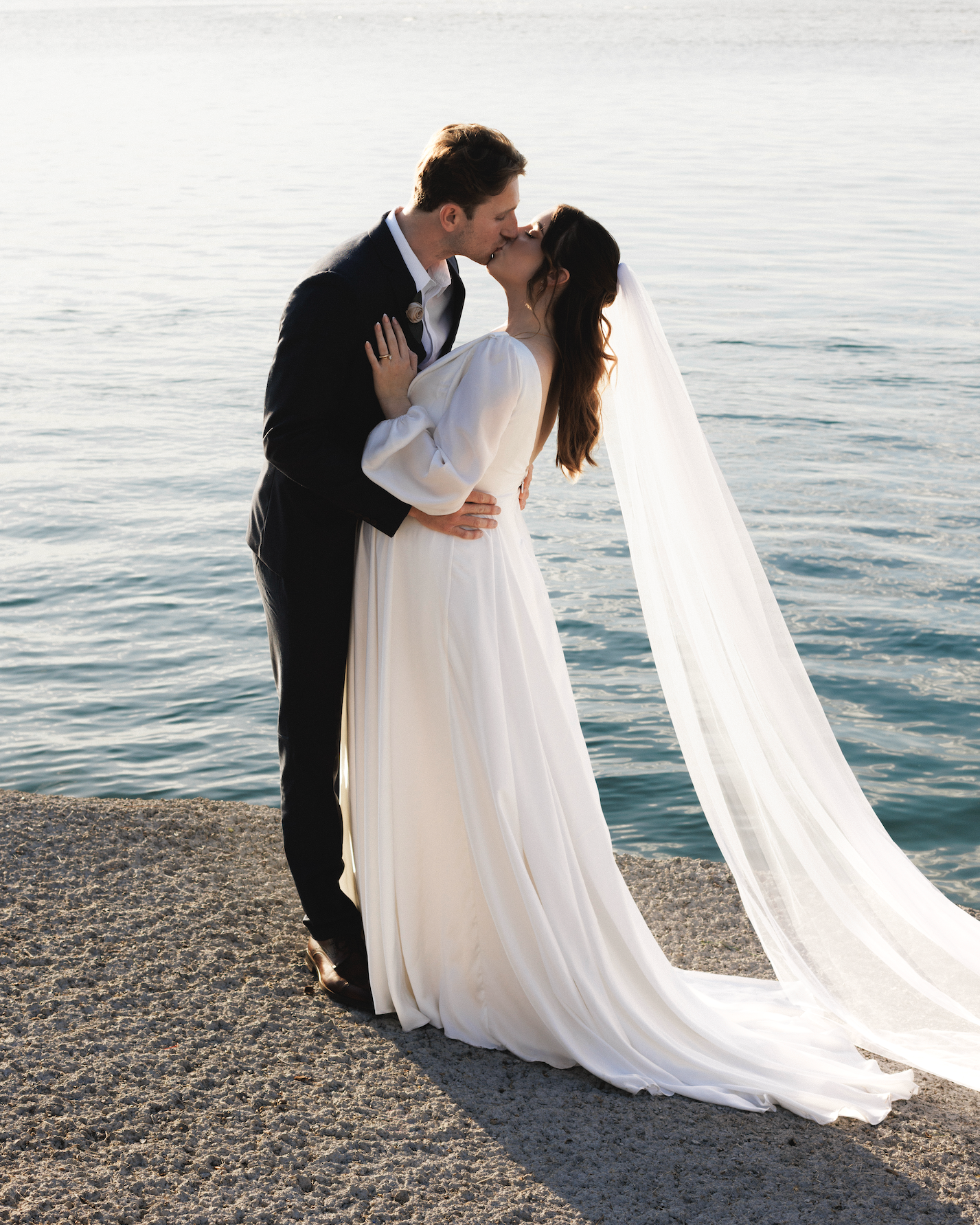 A newlywed couple kissing by a body of water, with the bride in a white wedding gown and veil, and the groom in a dark suit, standing on a textured concrete surface.