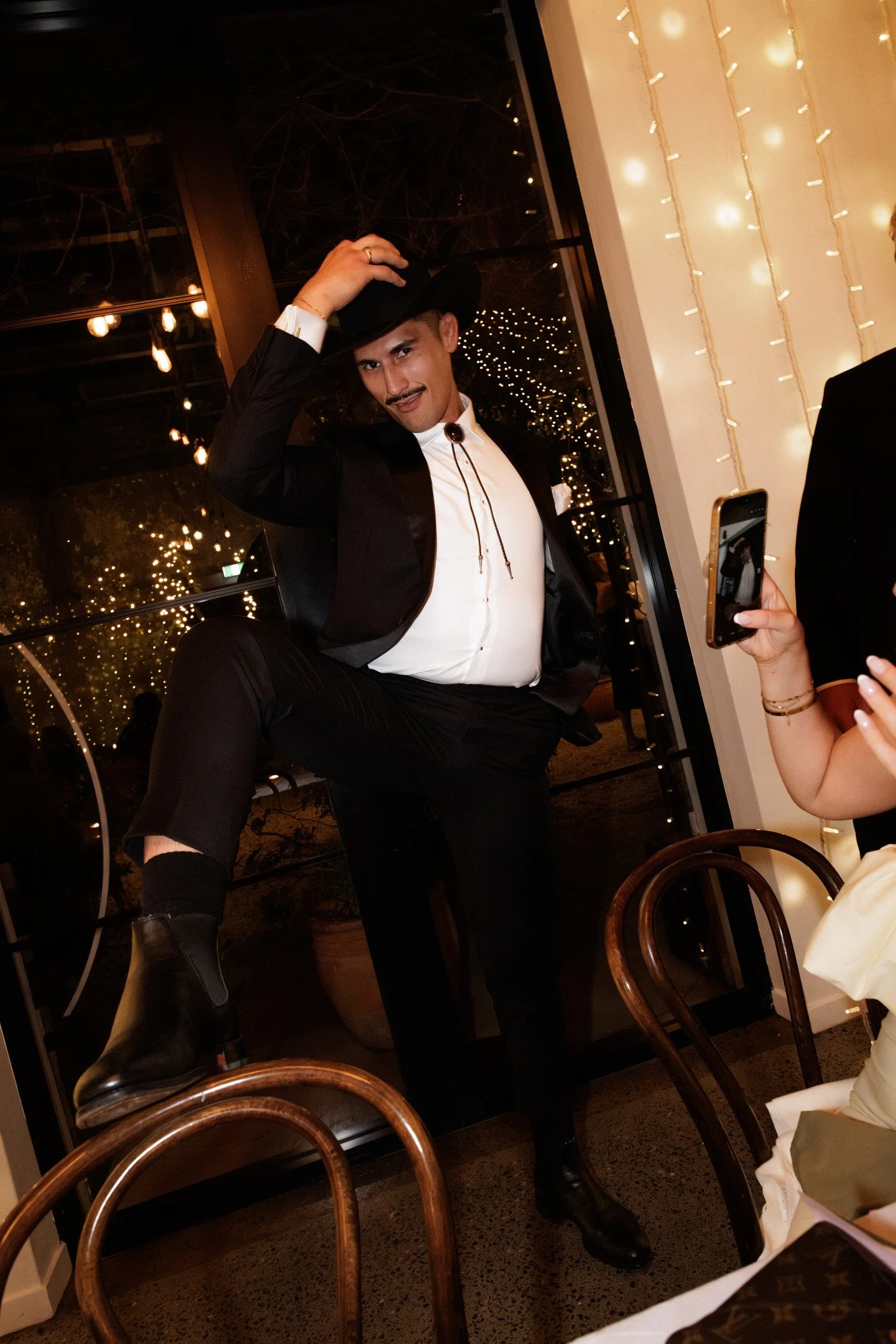 A man dressed in a tuxedo with a bolo tie, black hat, and boots is confidently posing inside a dimly lit restaurant with warm string lights. Someone is taking a photo of him using a smartphone.