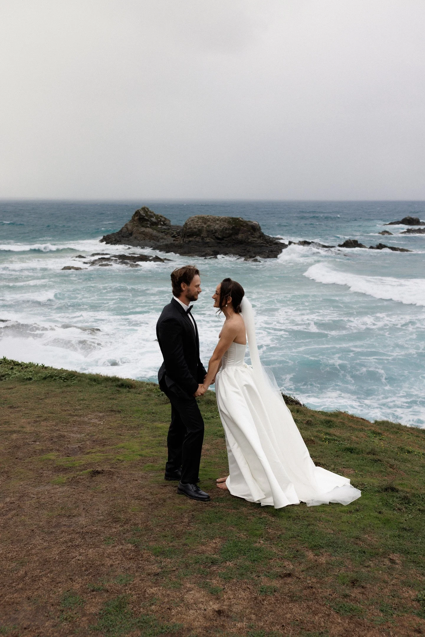 A bride and groom stand holding hands on a grassy area by the ocean, with waves crashing against rocks in the background. The groom wears a black tuxedo, and the bride wears a white strapless wedding gown and veil.
