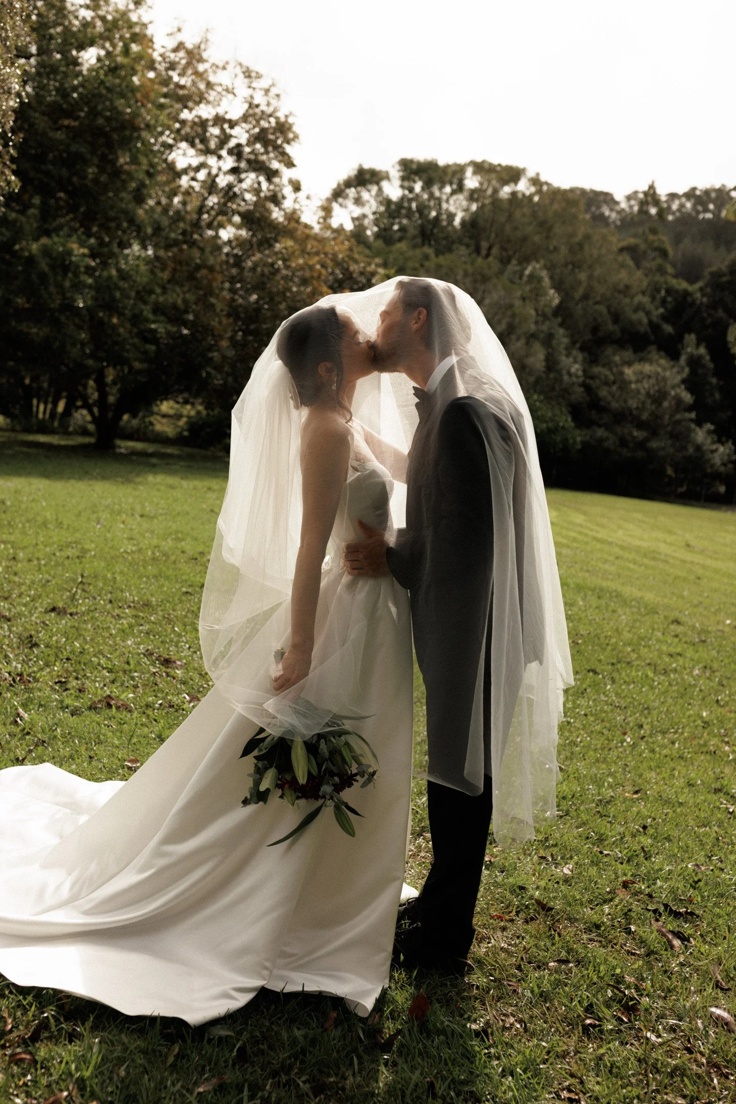 A bride and groom kissing under a wedding veil outdoors in a park with trees in the background.