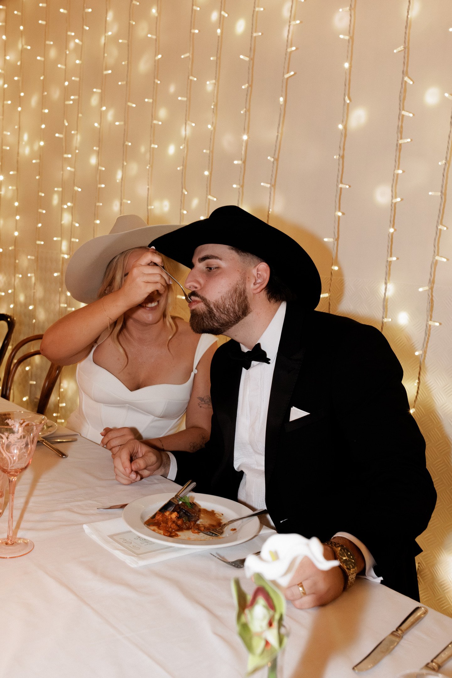 A woman in a white dress and tan hat feeds a man in a tuxedo and black cowboy hat at a wedding reception. They are sitting at a table with food, wine glasses, and flowers, with fairy lights in the background.