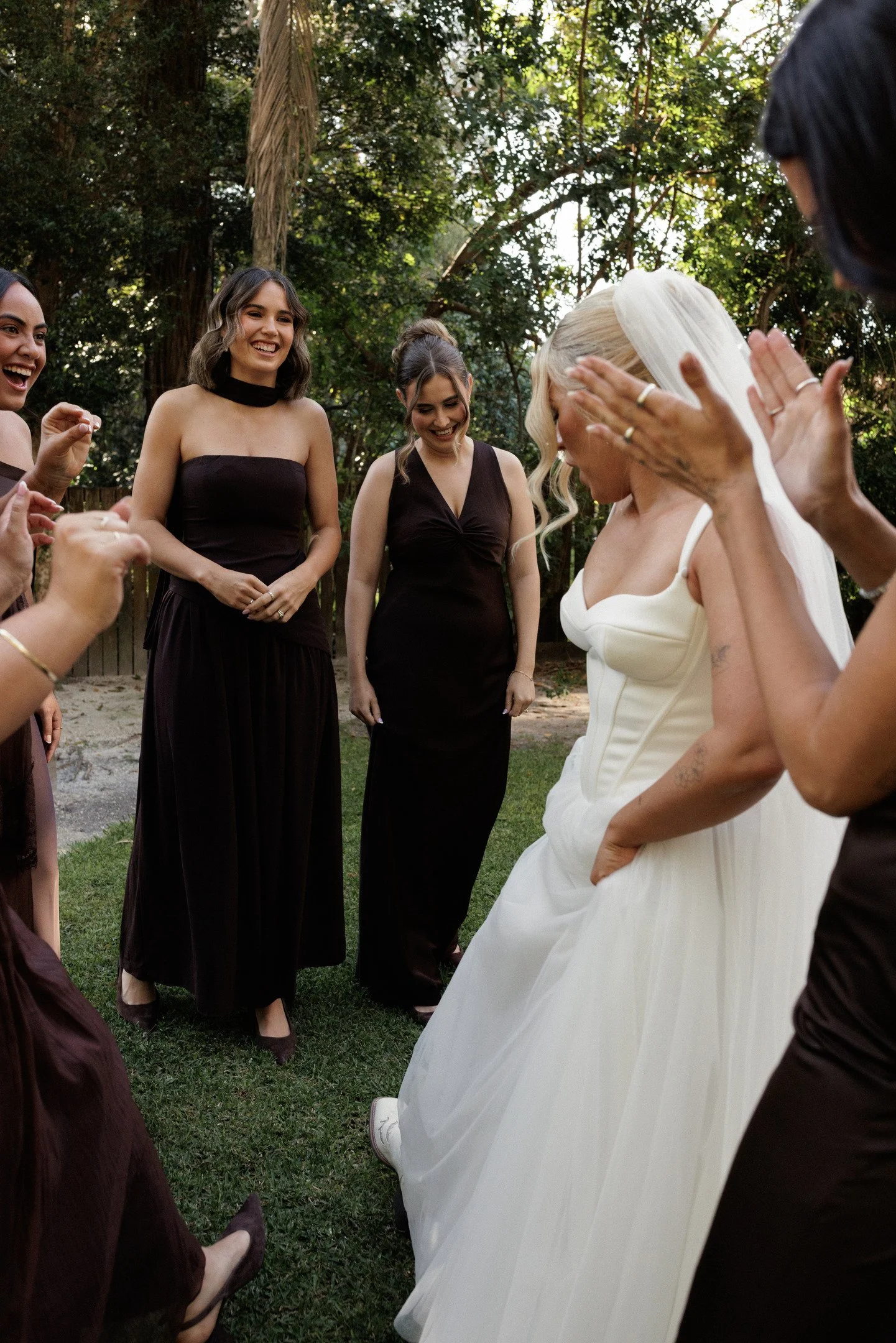 A bride in a white wedding dress is laughing and covering her face with her hands, surrounded by smiling women in matching black dresses in a garden setting.