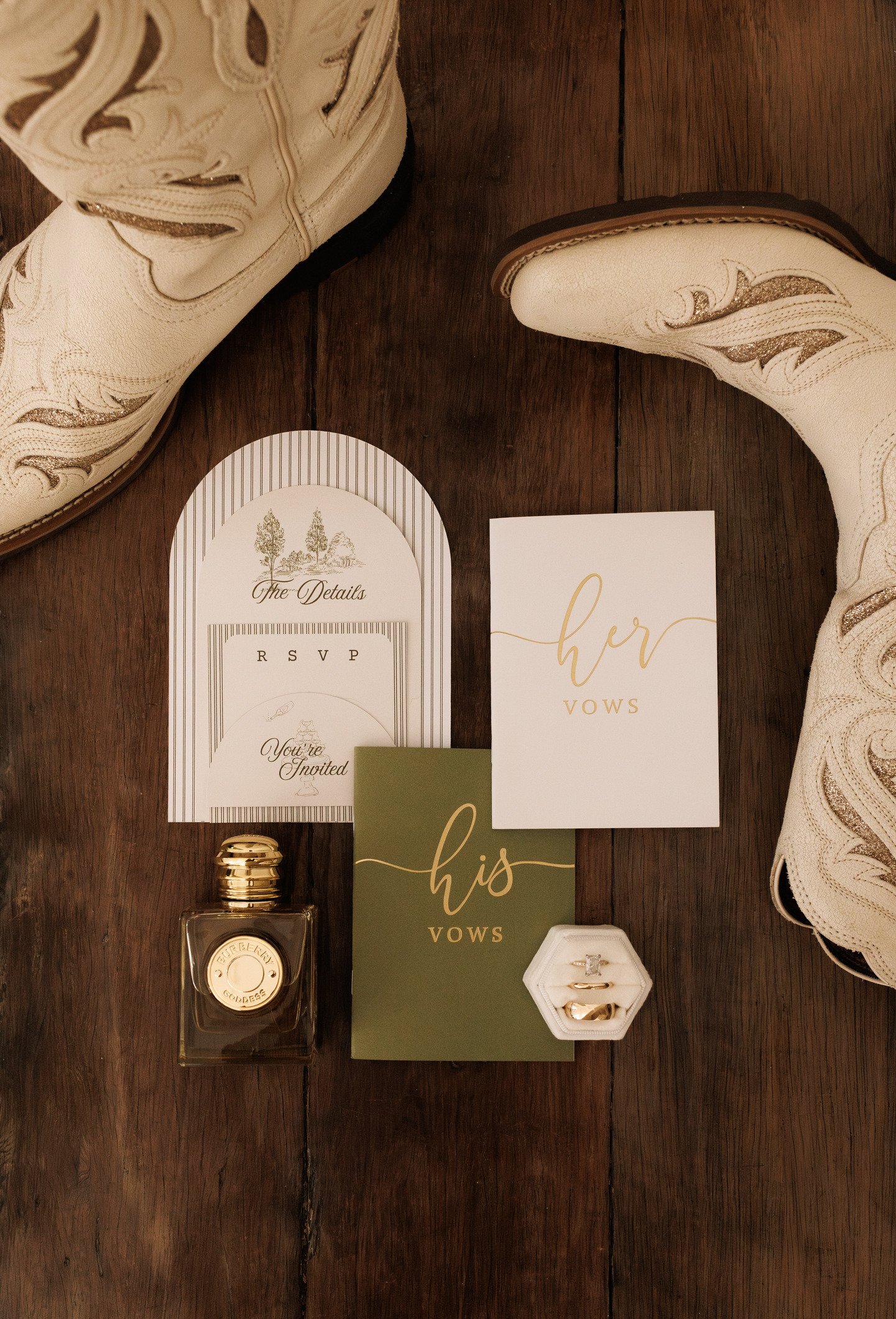 A pair of white cowboy boots with gold decorative stitching, wedding rings in a white ring box, wedding vow cards reading 'here' and 'his' with gold lettering, a gold perfume bottle, and an RSVP card with a tree illustration on dark wooden surface.