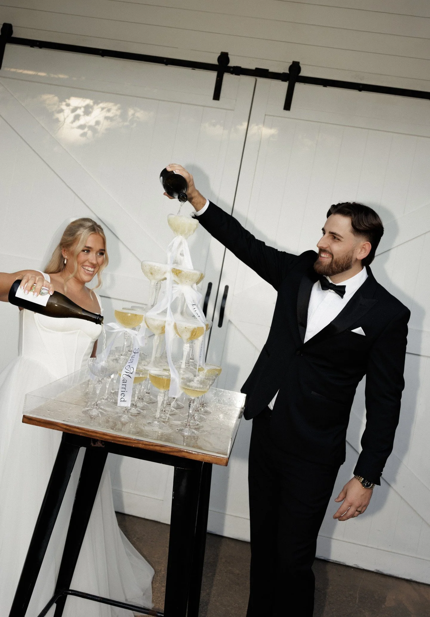 A man and woman dressed in formal attire celebrating at a party, pouring liquid into a champagne tower made of stacked glasses with white ribbons and a note reading 'Best Married' on the table.