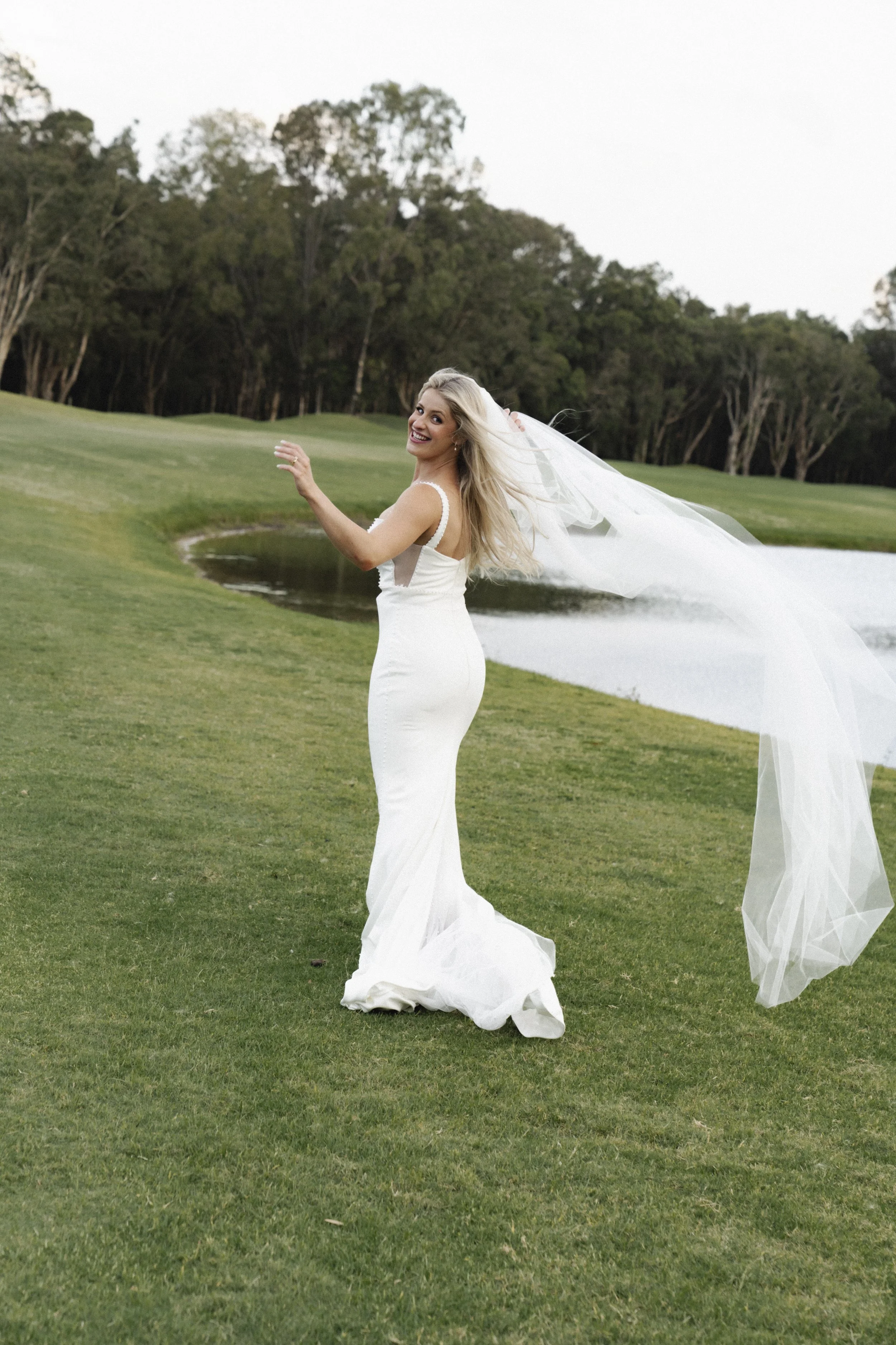 Bride in a white wedding dress walking on a grassy field by a pond, with trees in the background and a long veil flowing behind - Elevated Weddings shot on 35mm film