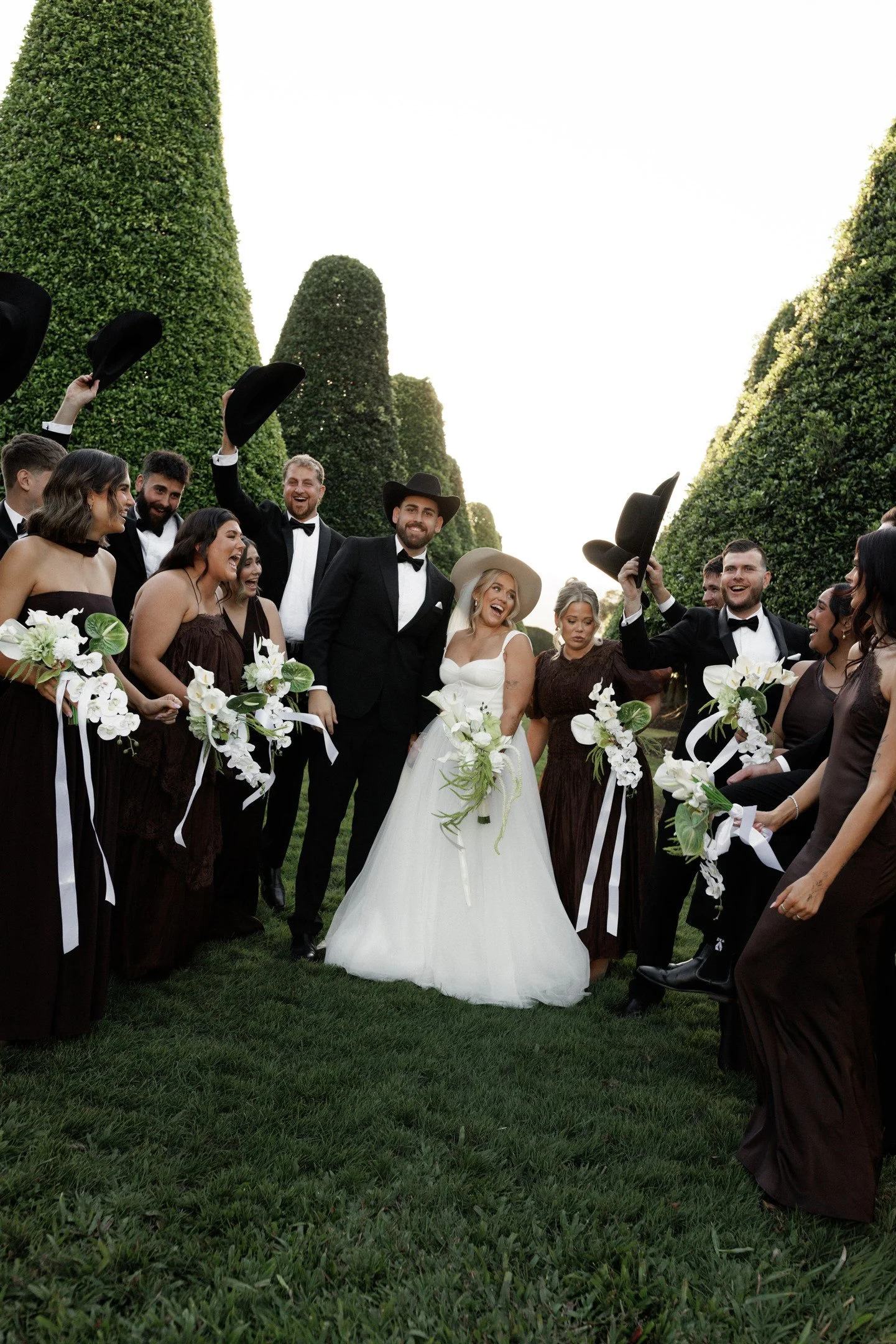 A wedding celebration outdoors with a bride in a white gown and guests dressed in black and white, smiling and holding hats in the air, surrounded by tall, lush green trees.