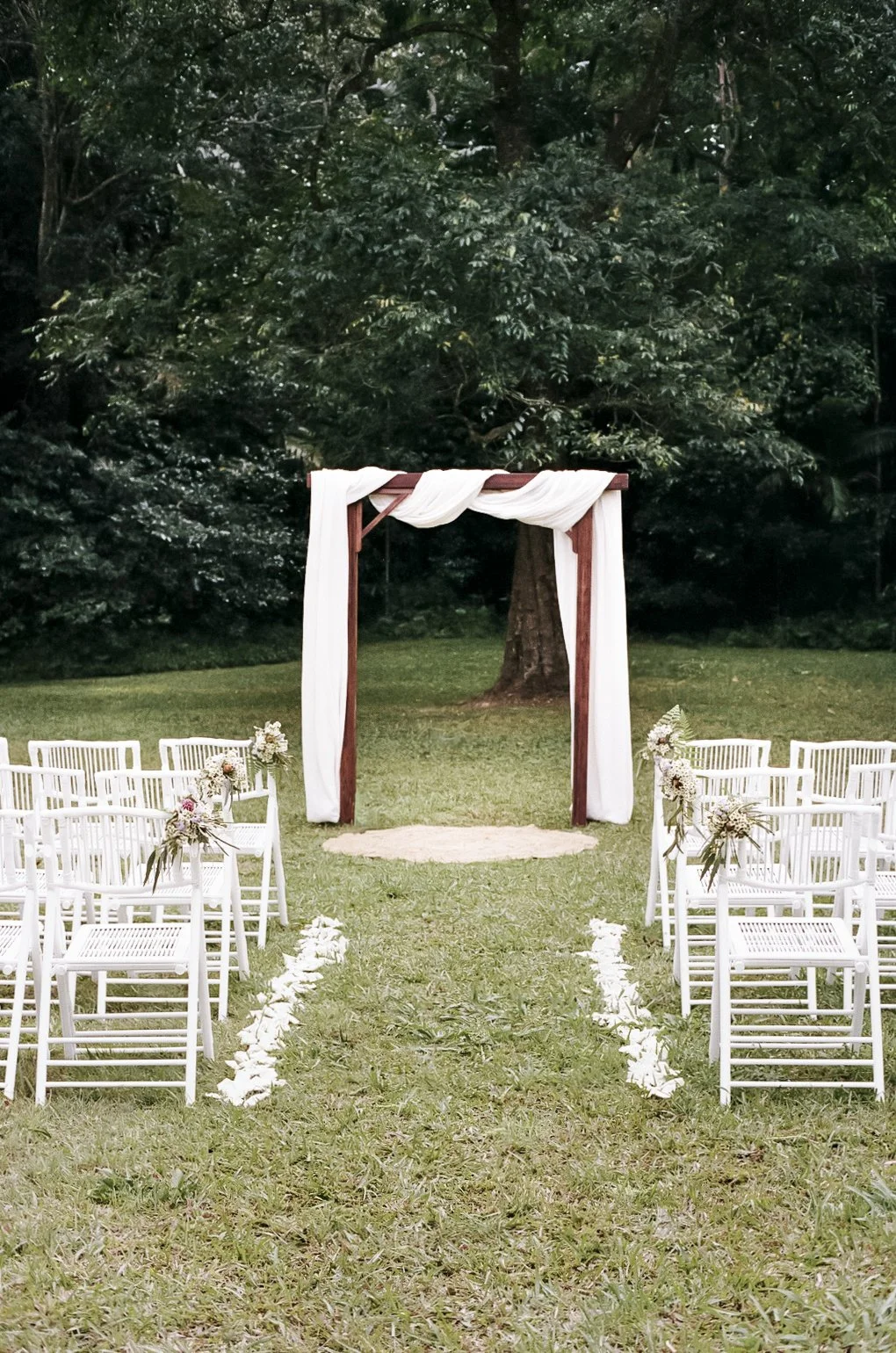 Outdoor wedding ceremony setup with white chairs decorated with flowers, facing a wooden arch draped with white fabric, on a grassy area surrounded by trees.