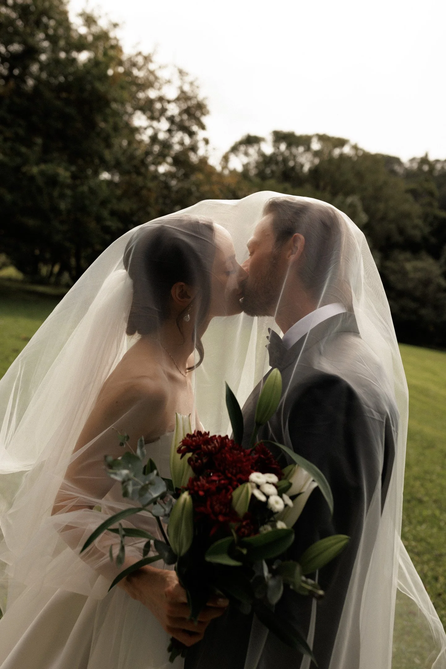 A bride and groom sharing a kiss under a veil during their outdoor wedding, holding a bouquet of flowers.