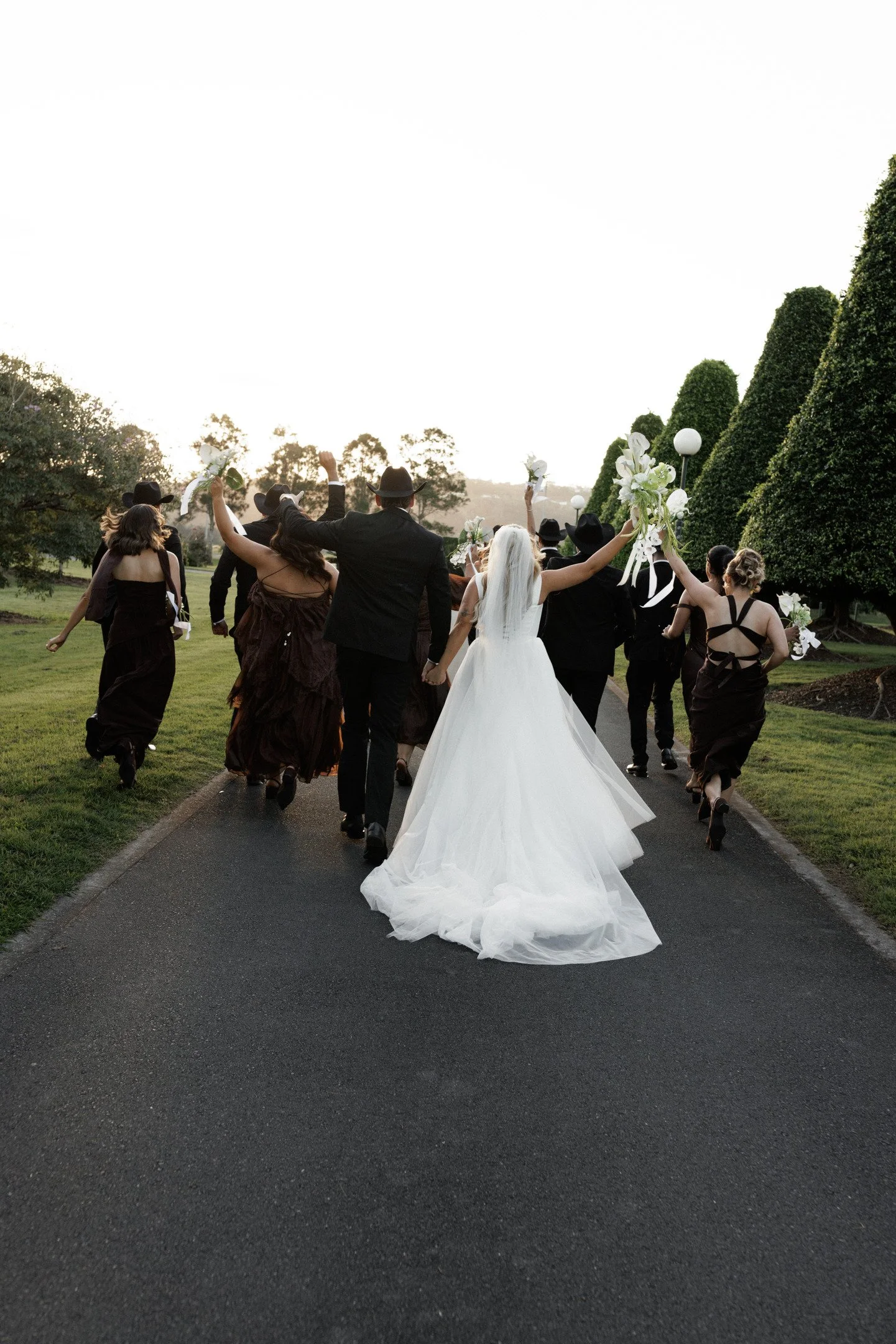 A bride and groom walking down a paved path surrounded by friends and family, everyone holding flowers and raising their hands in celebration, during a wedding ceremony outdoors at sunset.