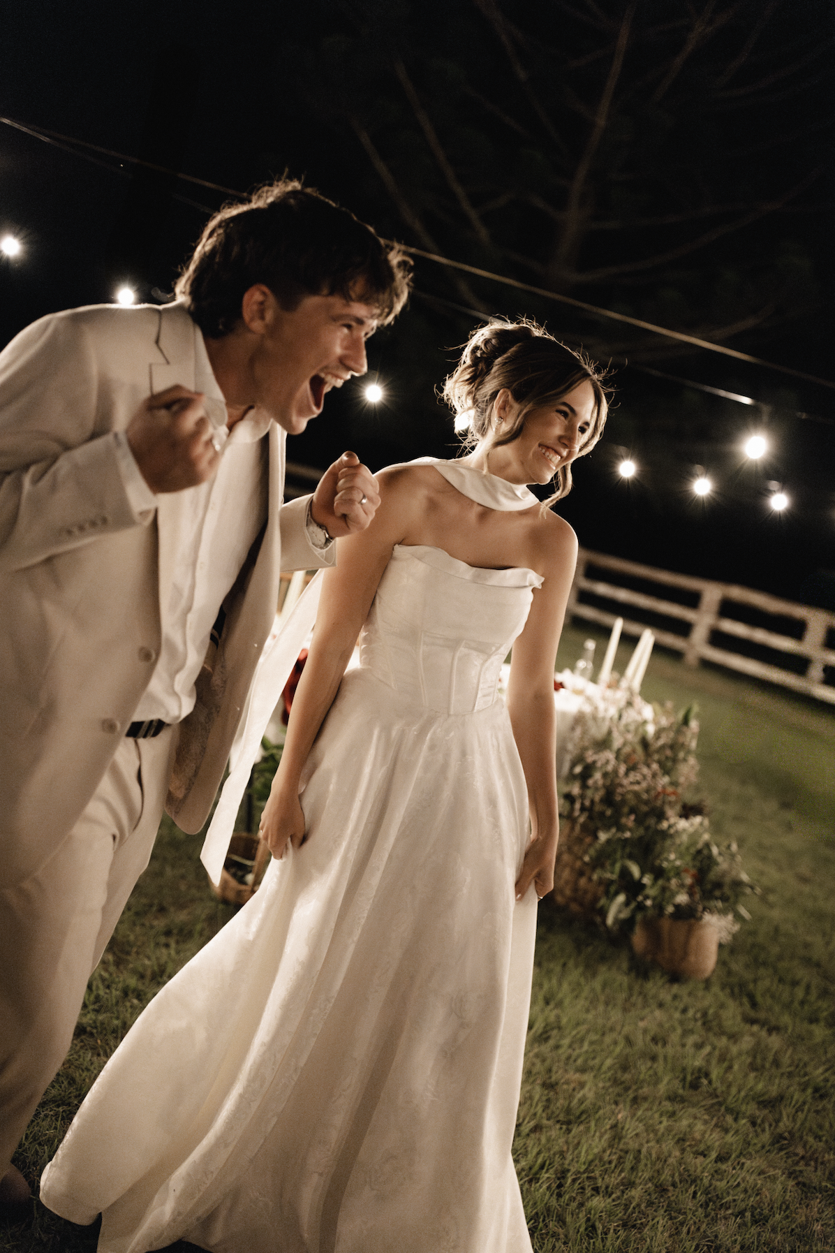A couple in wedding attire celebrating outdoors at night, with strings of lights overhead and a decorated table in the background.
