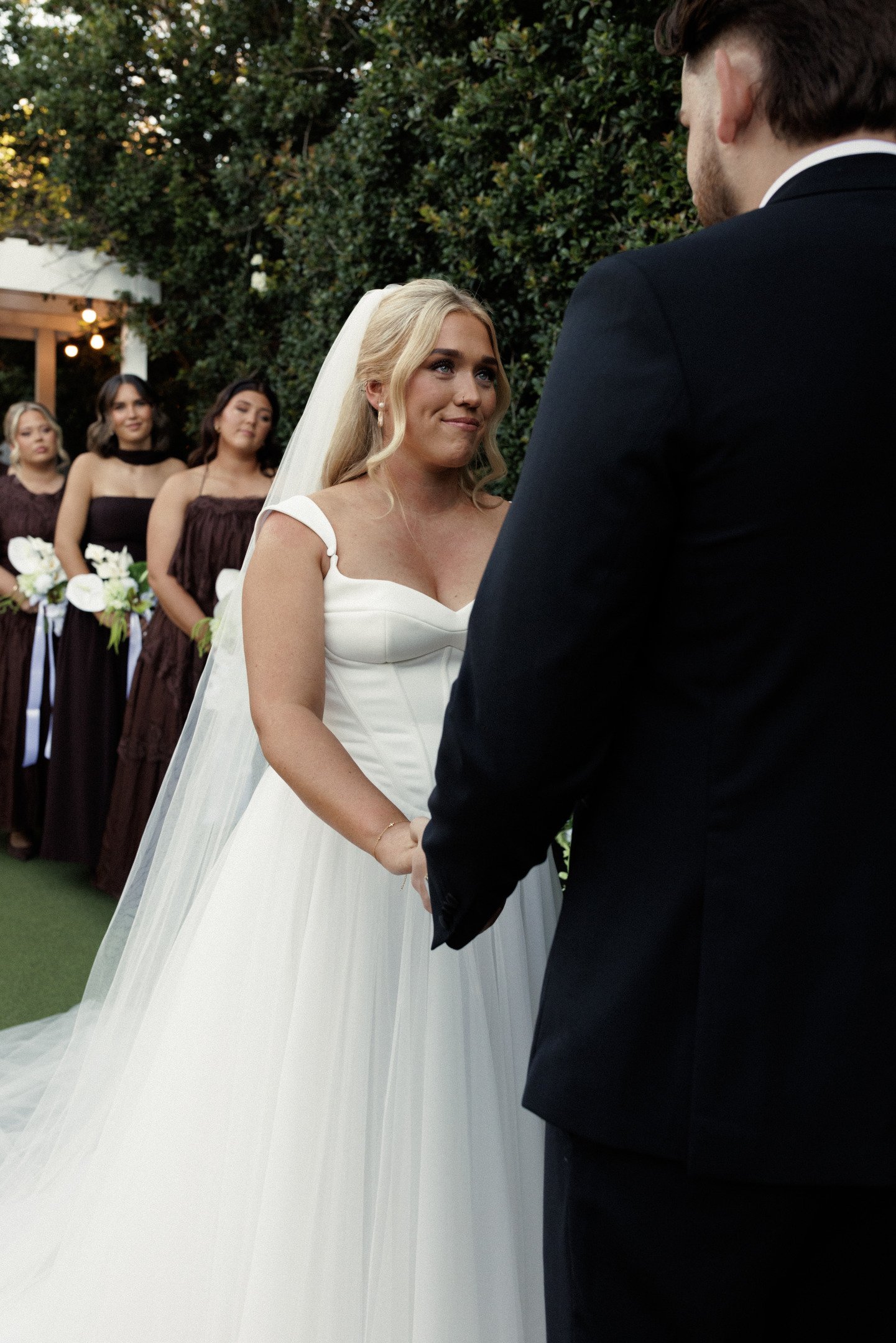 A bride and groom exchange vows during an outdoor wedding ceremony, with bridesmaids in dark dresses standing in the background.