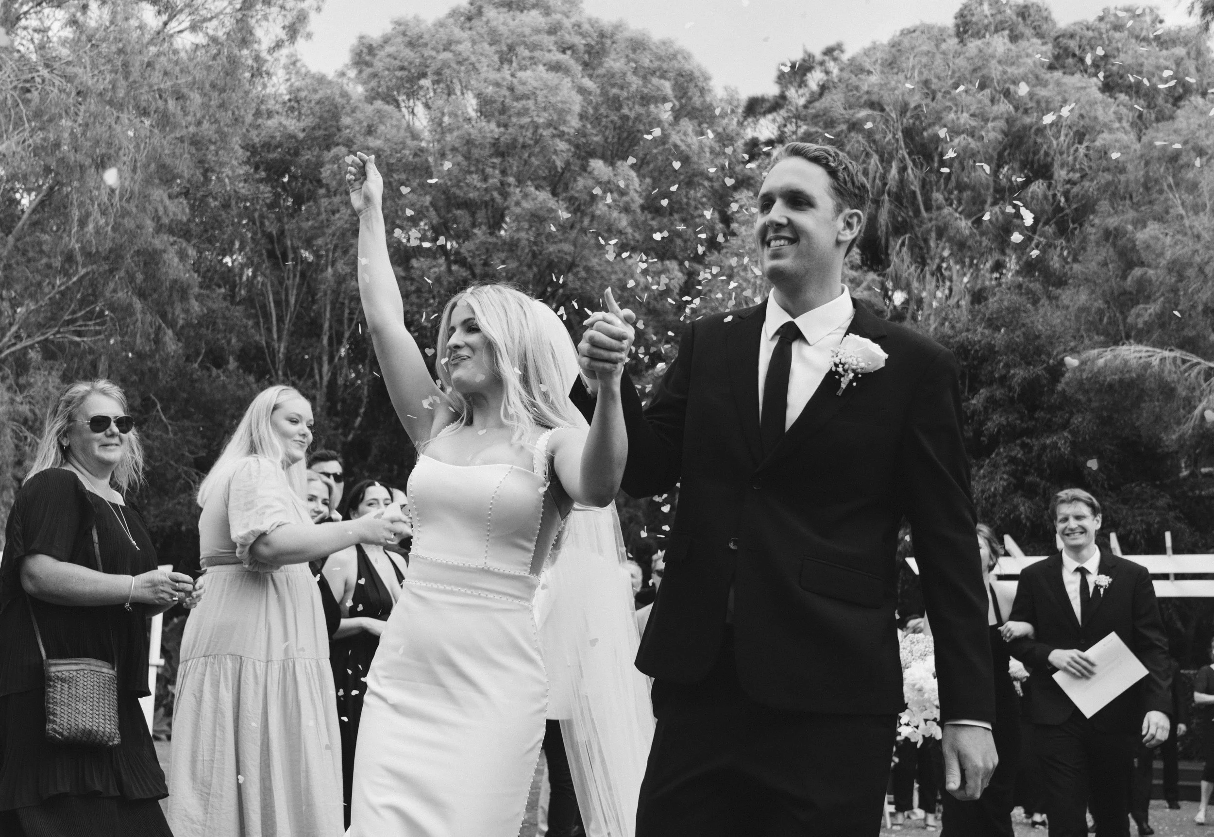 A black and white photo of a wedding celebration with a bride and groom smiling and holding hands, surrounded by friends and family outdoors. The bride is waving while the groom holds her hand, with people of various ages watching and smiling.