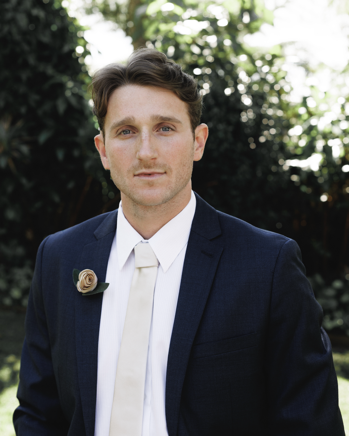 A young man in a navy suit with a white shirt and tie, wearing a beige rose boutonniere, standing outdoors with green foliage in the background.