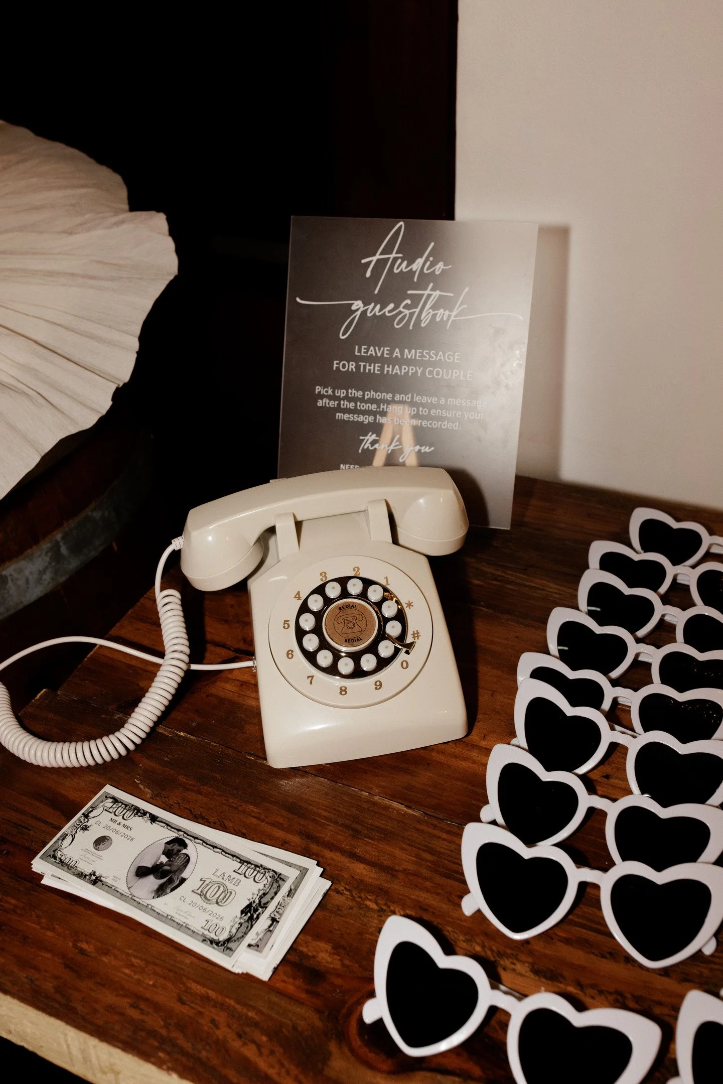 A vintage rotary phone on a wooden surface with a black and white sign next to it that says "Audio guestbook" and instructions to leave a message for a happy couple. There are also black heart-shaped cutouts on a white background and some stacks of U