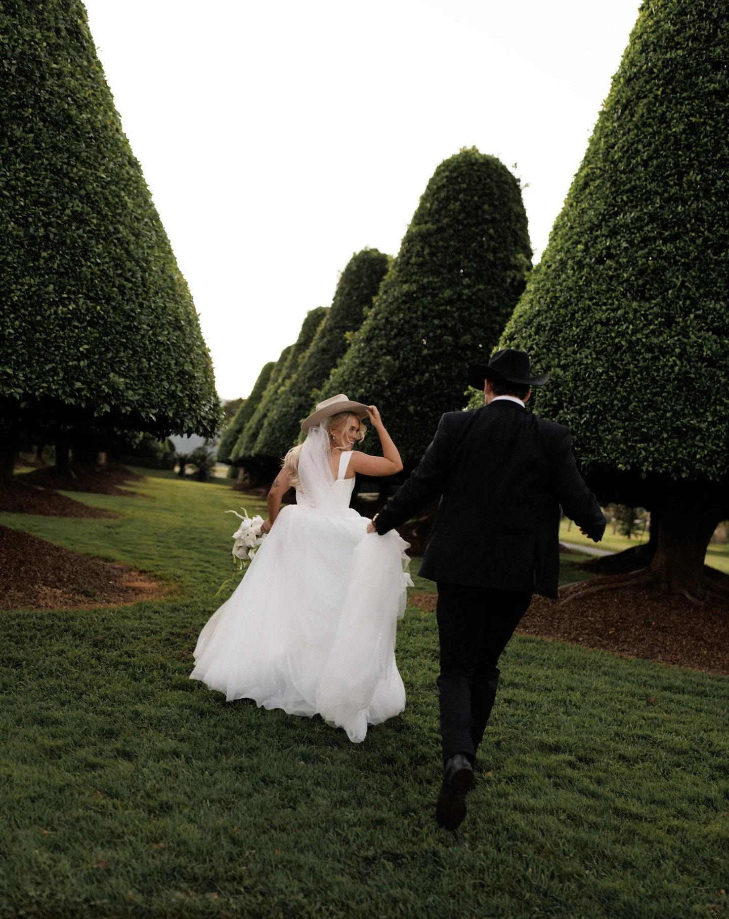 A bride and groom holding hands walking through a garden with large, manicured trees in the background, during what appears to be a wedding photo shoot.