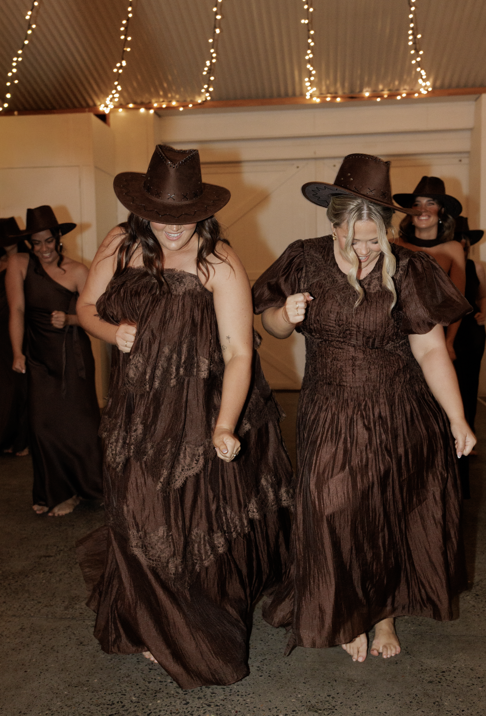 A group of women dancing together at a party or celebration, wearing brown dresses and cowboy hats, with string lights hanging from the ceiling.