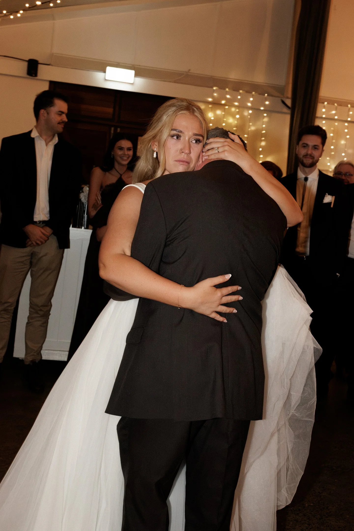 A bride and groom hugging during their wedding reception. The bride looks emotional, while guests in formal attire are smiling and watching in the background. The setting is decorated with string lights.