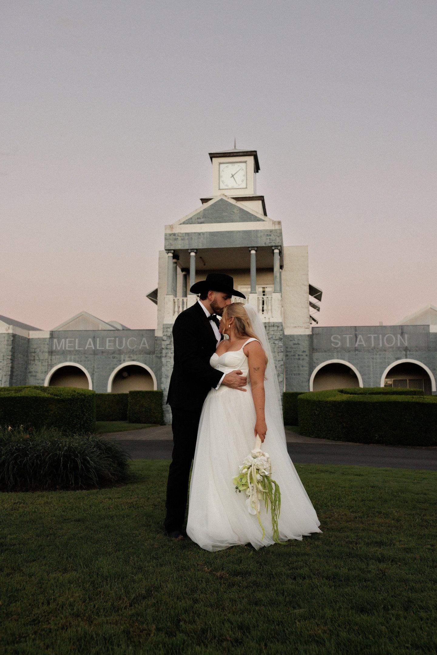 A bride and groom sharing a tender moment in front of a building with a clock tower and the words "Melaleuca Station" on the wall.