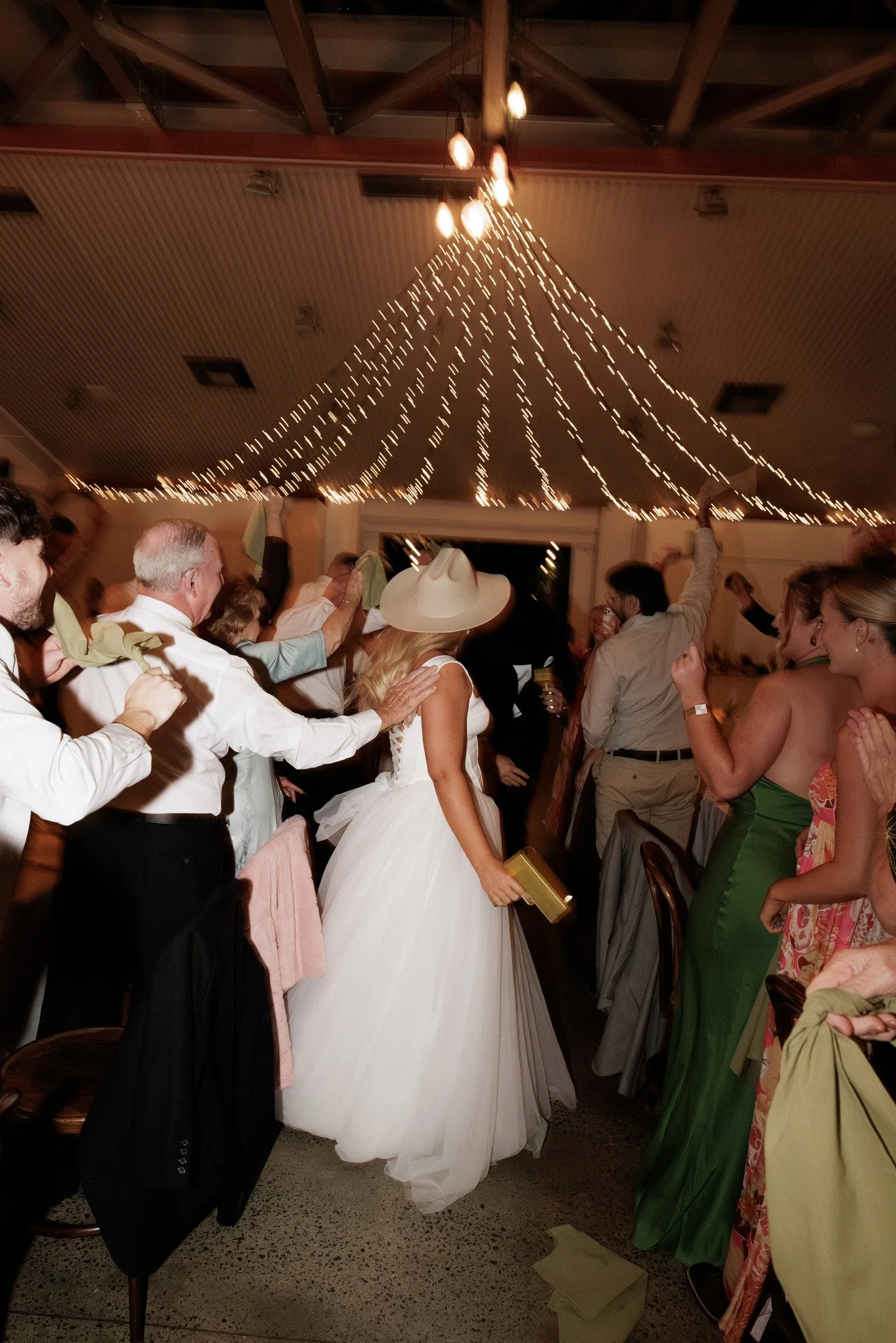 People dancing and celebrating at a wedding reception, with a ceiling decorated with string lights.