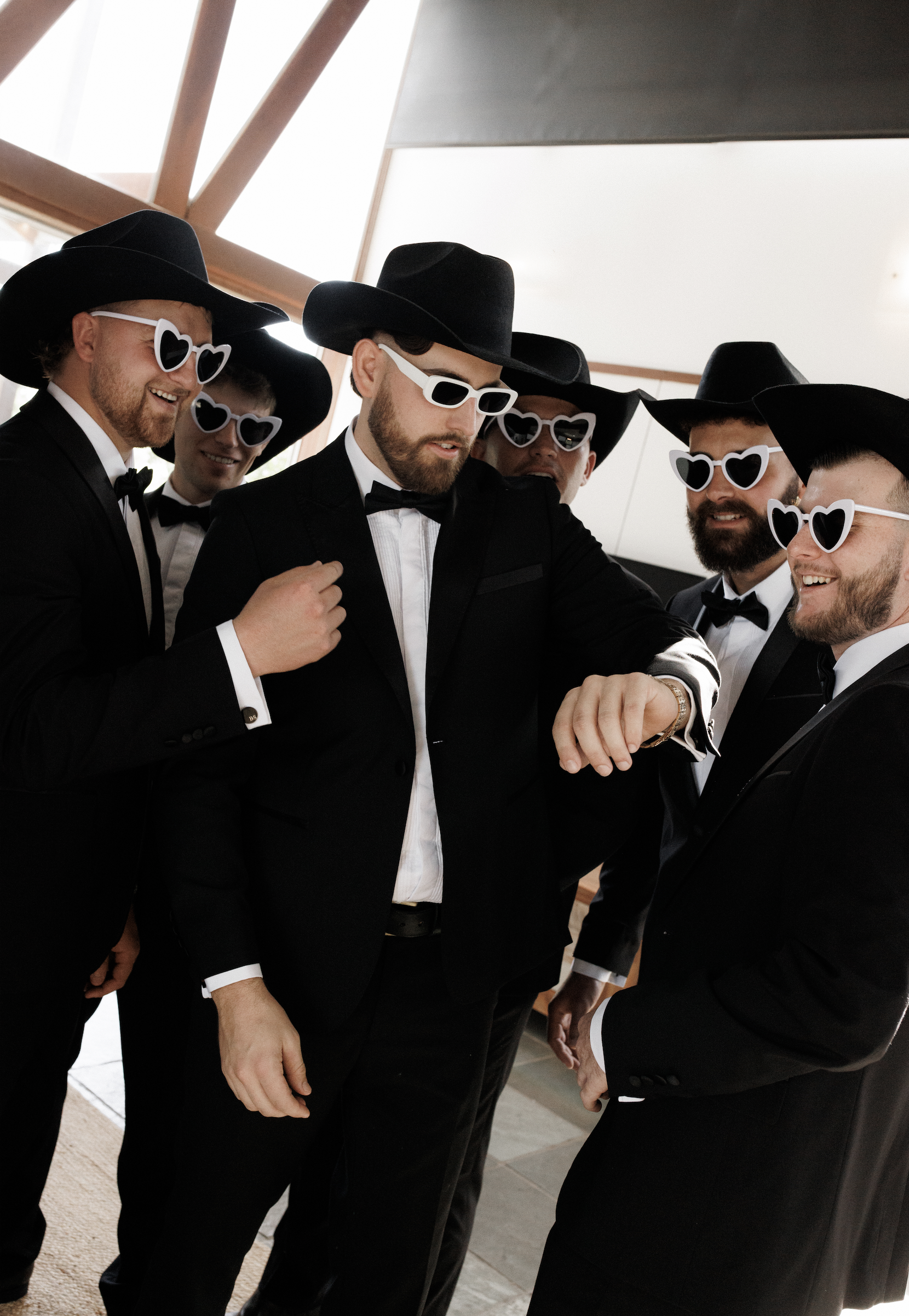 A group of six men dressed in black tuxedos and black hats, wearing white-framed sunglasses with heart-shaped lenses, gathered indoors, looking at a watch, smiling and joking around.