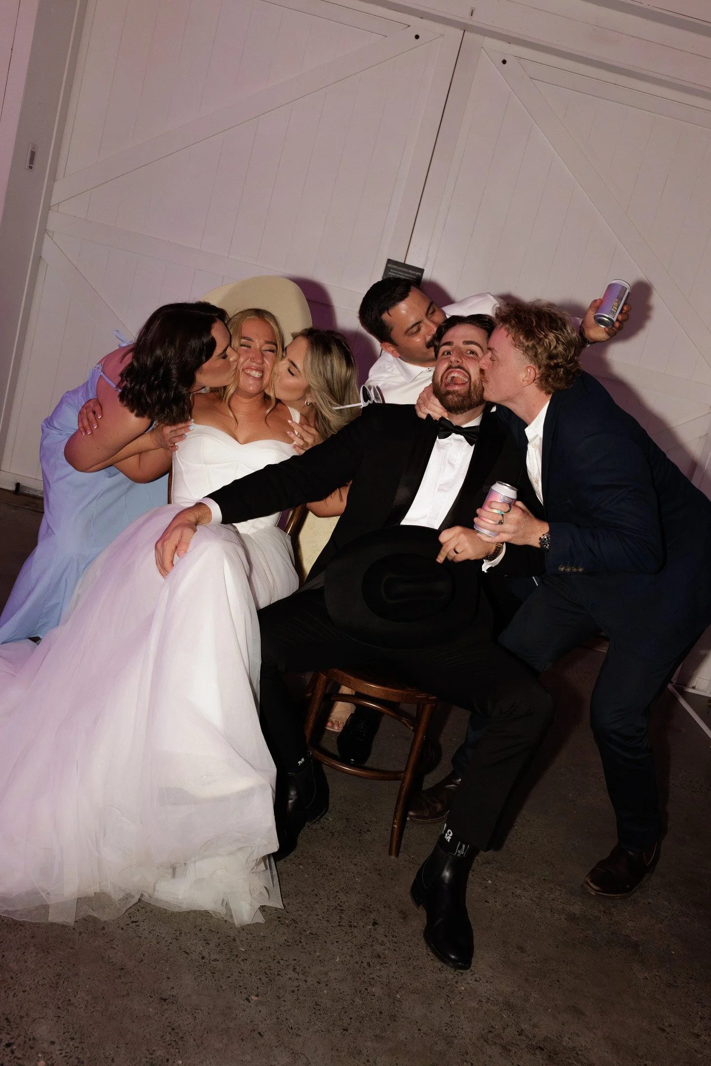 Group of five people, including a bride in a white wedding dress, celebrating and kissing at a wedding reception, with some holding drinks, all smiling and joyfully interacting.