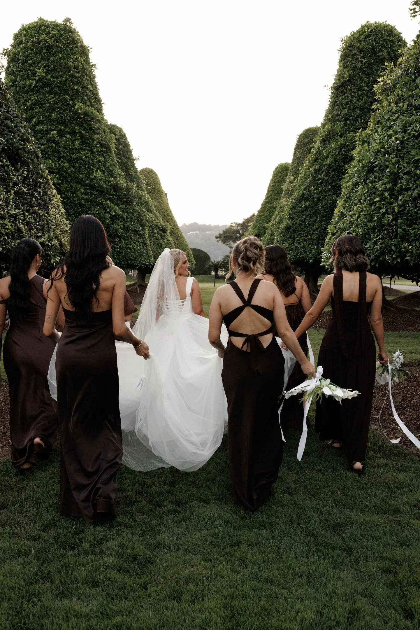 A bride in a white wedding dress holding hands with a group of women dressed in black or dark brown gowns, walking outdoors through a tree-lined pathway with trimmed trees and a grassy area.