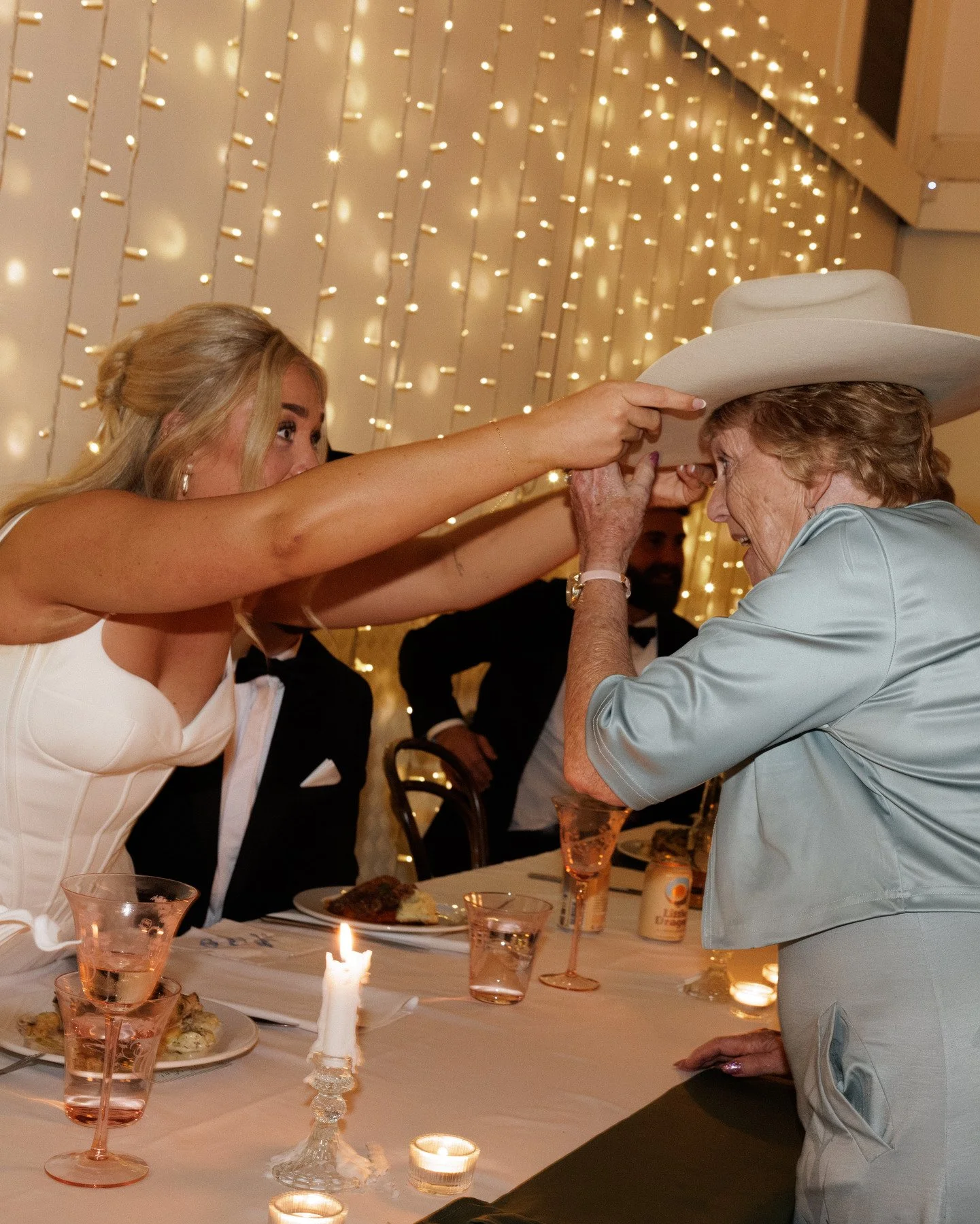 A young woman in a white dress is placing a hat on an elderly woman with short curly hair, wearing a light-colored outfit, at a celebration or party. The background features string lights, and a table is set with wine glasses, plates of food, candles