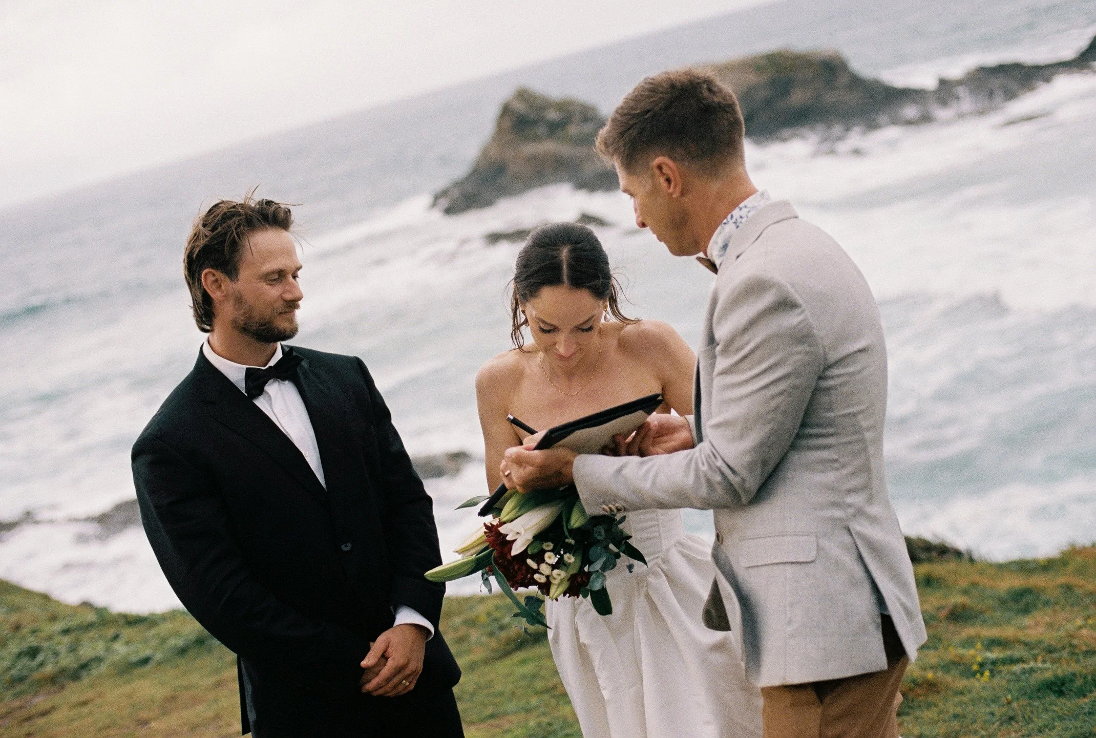 A wedding ceremony on a beach with three individuals: a man in a black tuxedo, a woman in a white dress holding a bouquet, and a man in a light gray suit officiating, with ocean waves and rocks in the background.