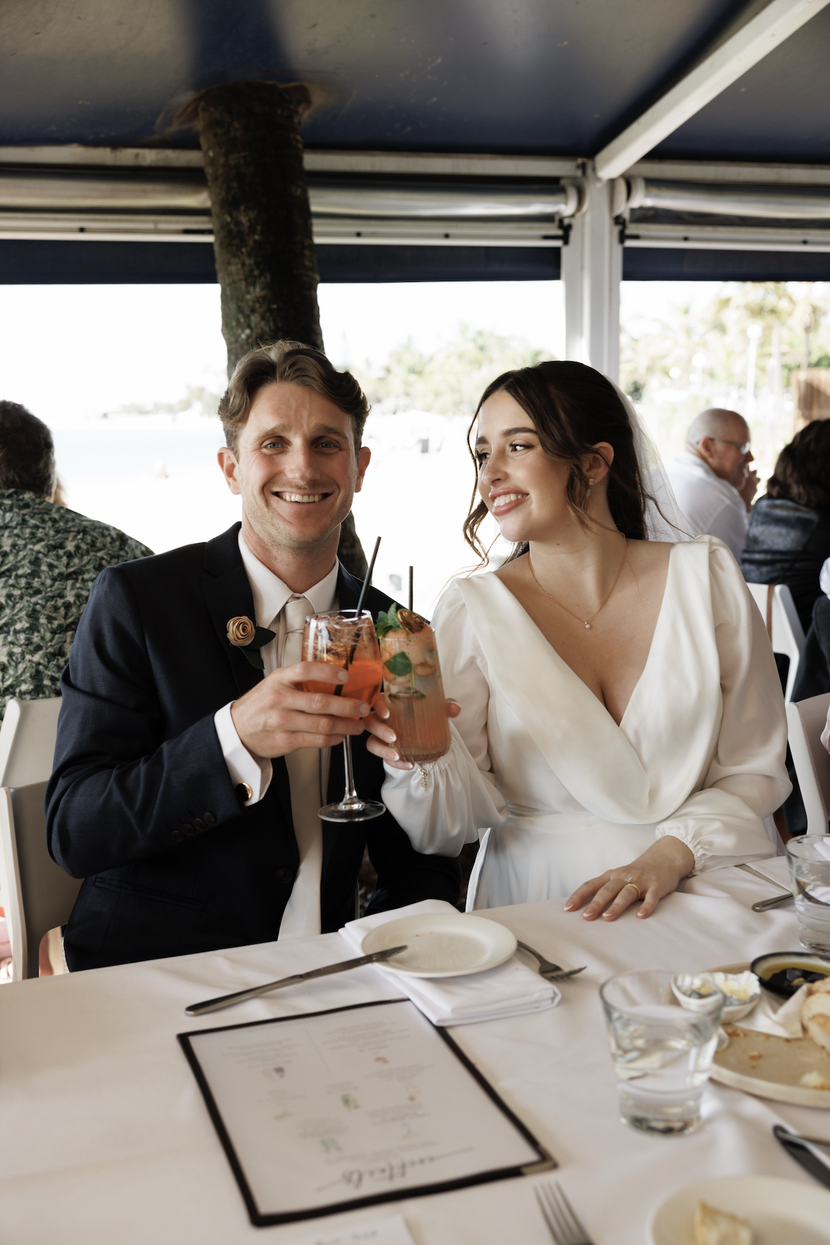 A bride and groom at their wedding reception enjoying drinks together, with other guests seated at the table.