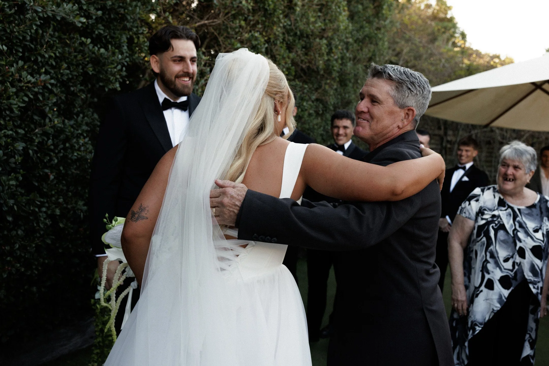 An outdoor wedding scene where a bride in a white wedding dress and veil embraces an older man, likely her father, while smiling. In the background, a groom in a black tuxedo with a bow tie and several guests, some in tuxedos and some in patterned dr
