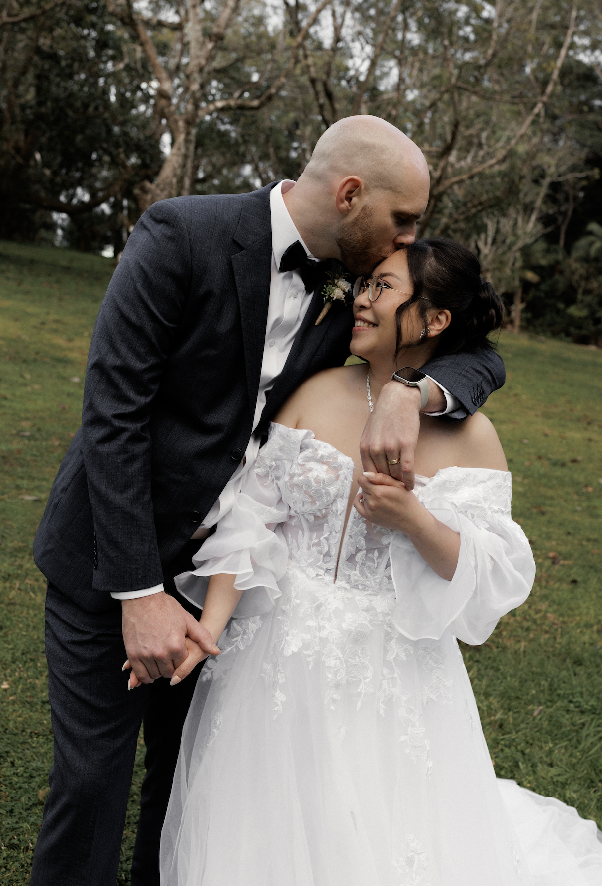 A newlywed couple, a man in a tuxedo with a bald head and beard kissing a woman in a white wedding dress with glasses, outdoors in a park with trees.