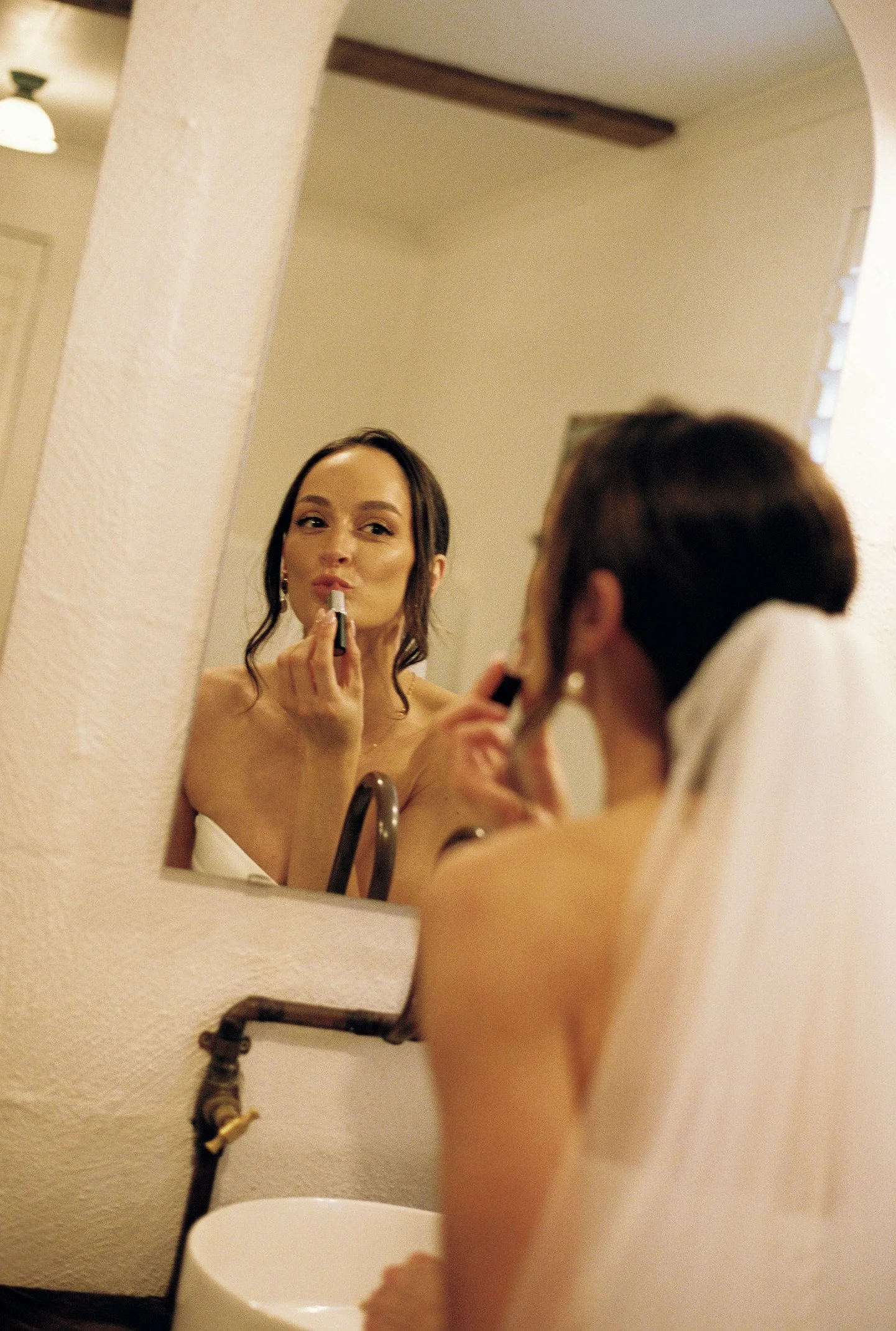 A woman with dark hair applying lipstick while looking into a mirror in a bathroom.