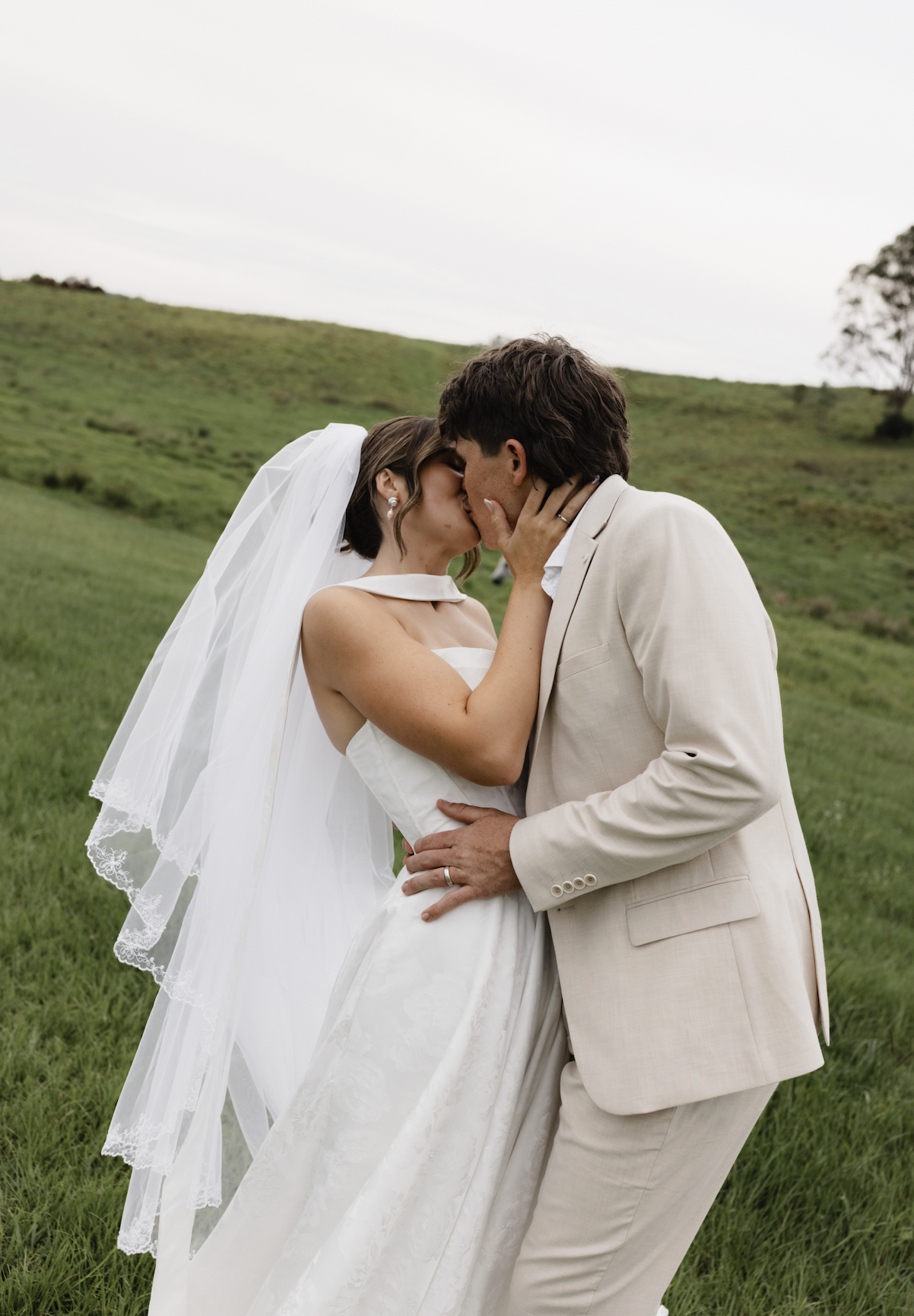 A bride and groom kissing outdoors in a grassy field with hills in the background, dressed in wedding attire.