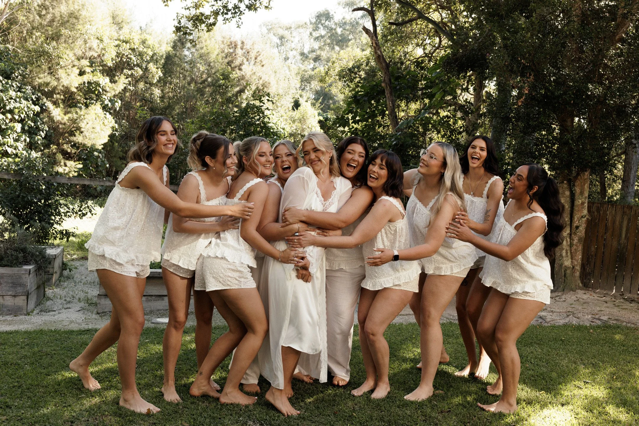 A group of women in white pajamas hugging and laughing outdoors in a green yard with trees.