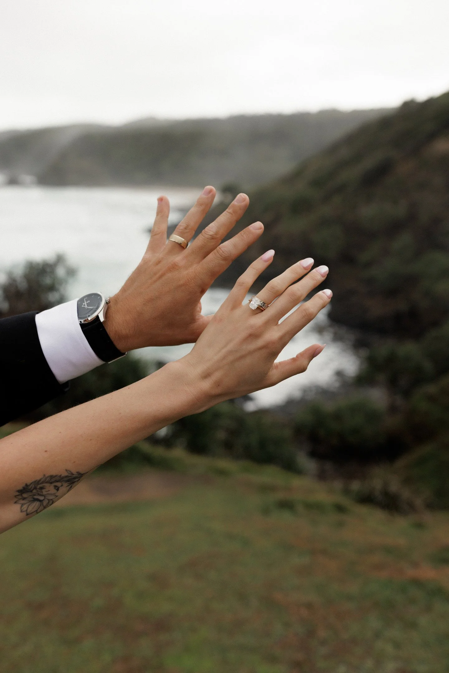 Four hands with rings and a watch in front of a scenic landscape with water and hills, outdoors during daytime.