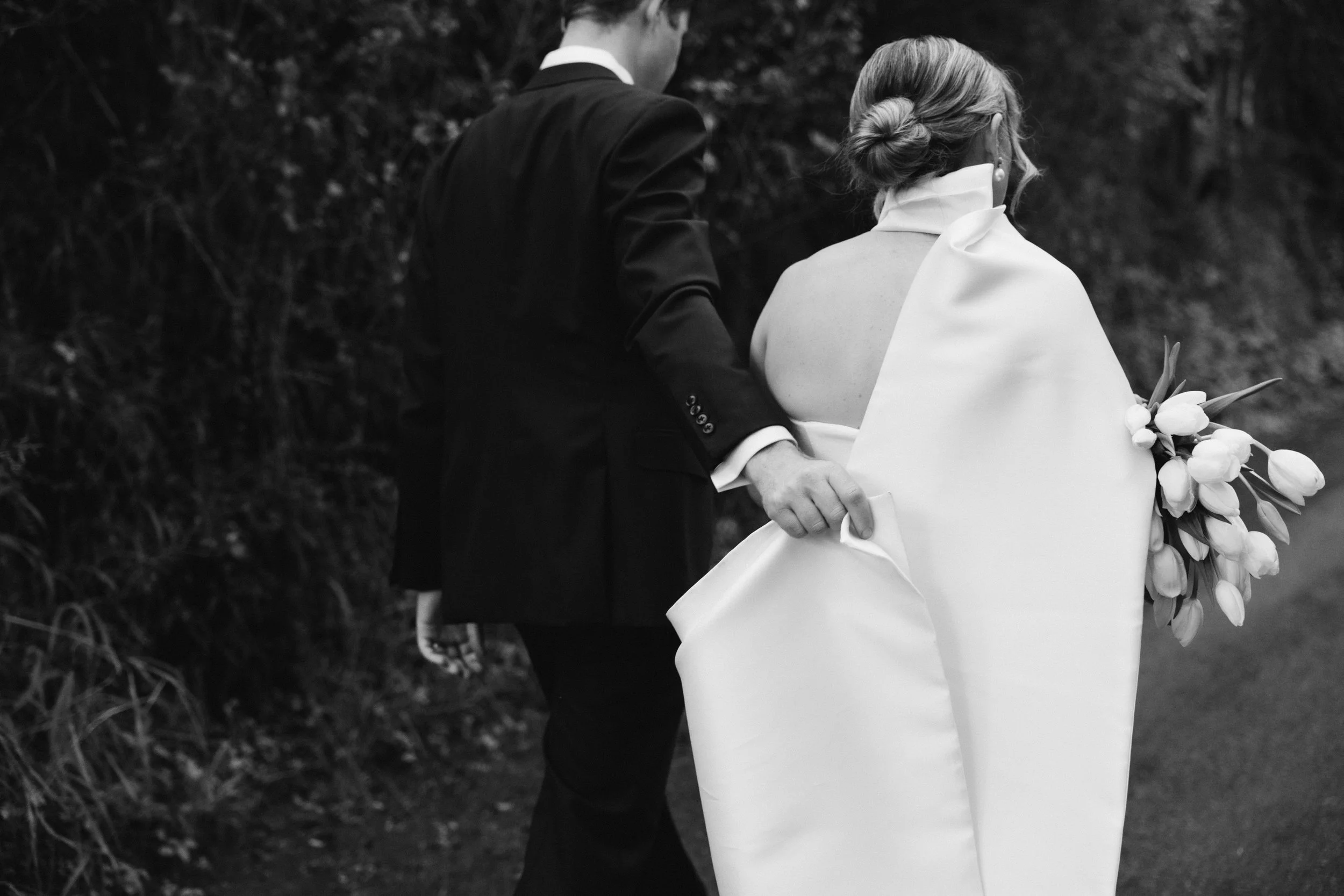 A black-and-white photo of a bride and groom walking outdoors. The groom is in a dark suit and the bride is in a light dress with a bouquet of tulips, with her hair styled in an updo.