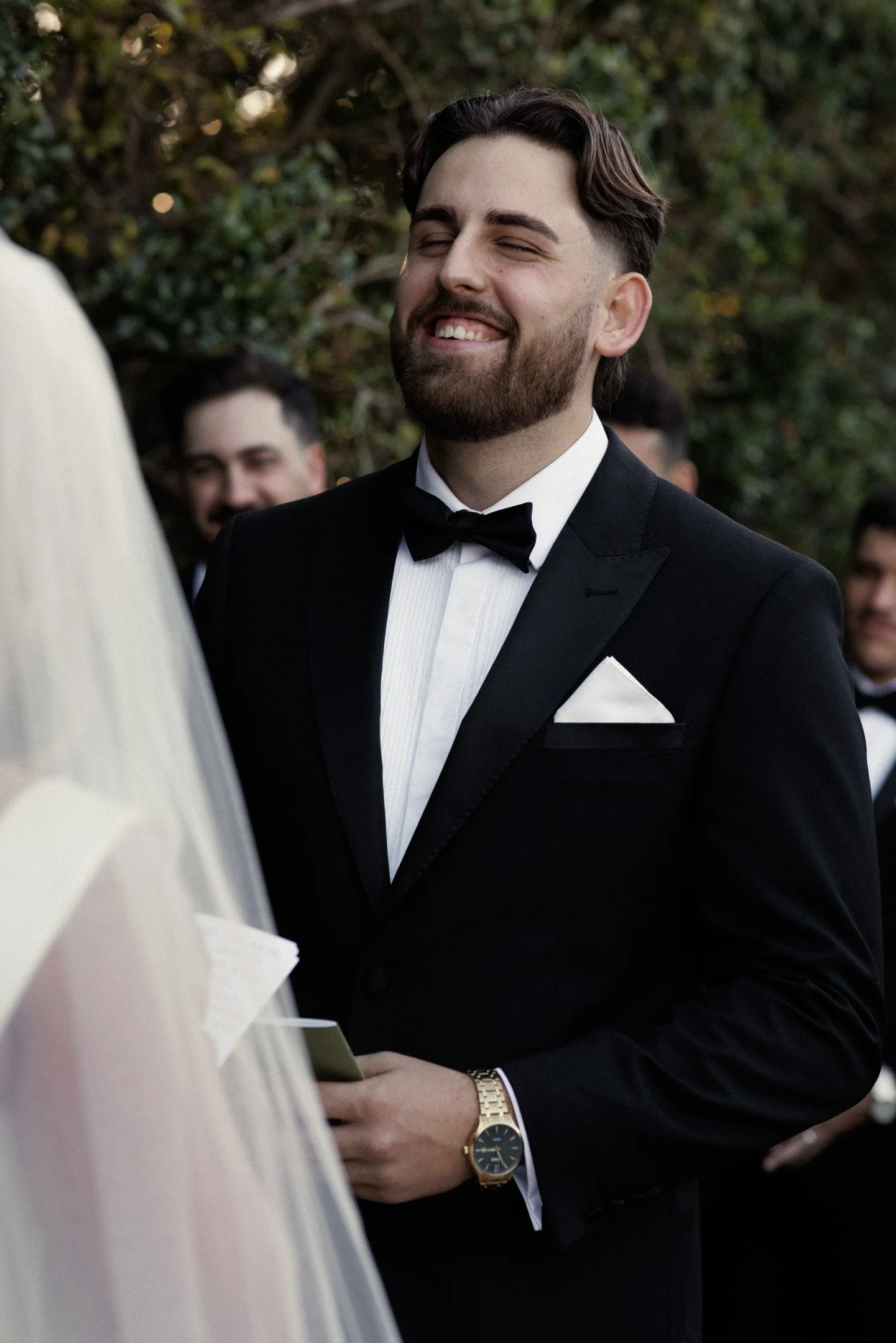 A groom in a black tuxedo with a bow tie smiling at a wedding ceremony, with guests and greenery in the background.