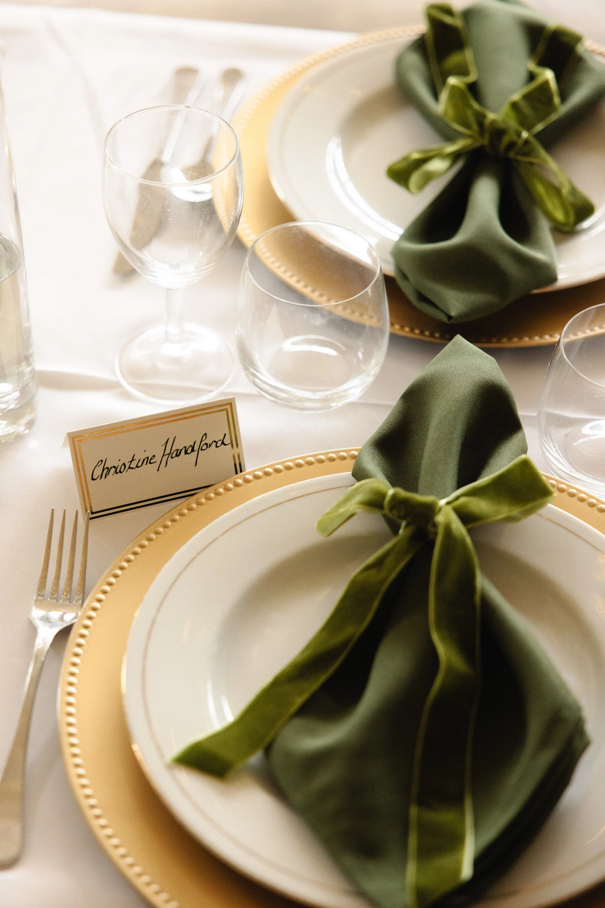 Elegant table setting with white plates, gold-rimmed chargers, green cloth napkins tied with green fabric, clear wine glasses, and a place card with the name 'Christine Haralord'