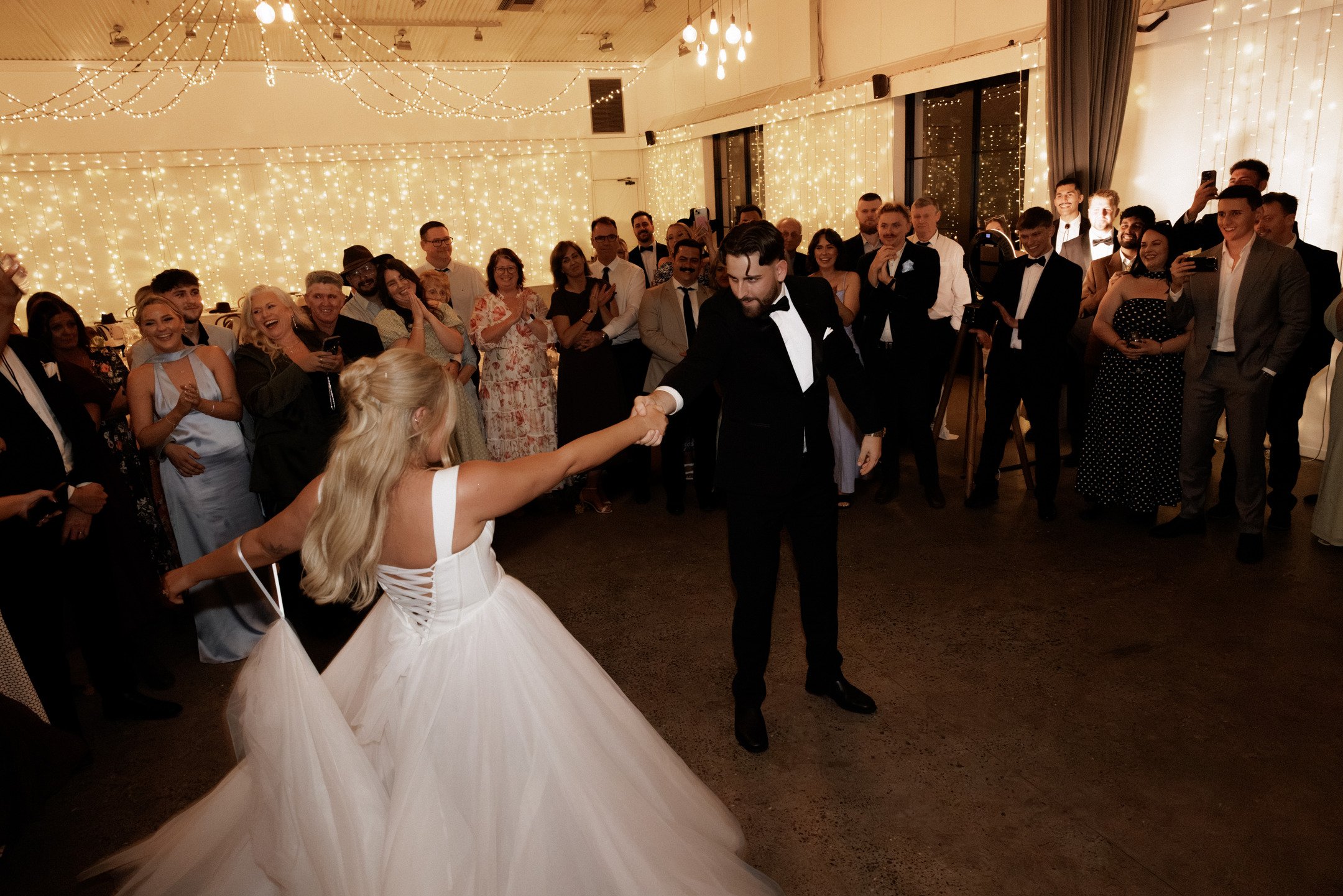 A wedding reception with guests watching the bride and groom dance, decorated with string lights hanging from the ceiling.