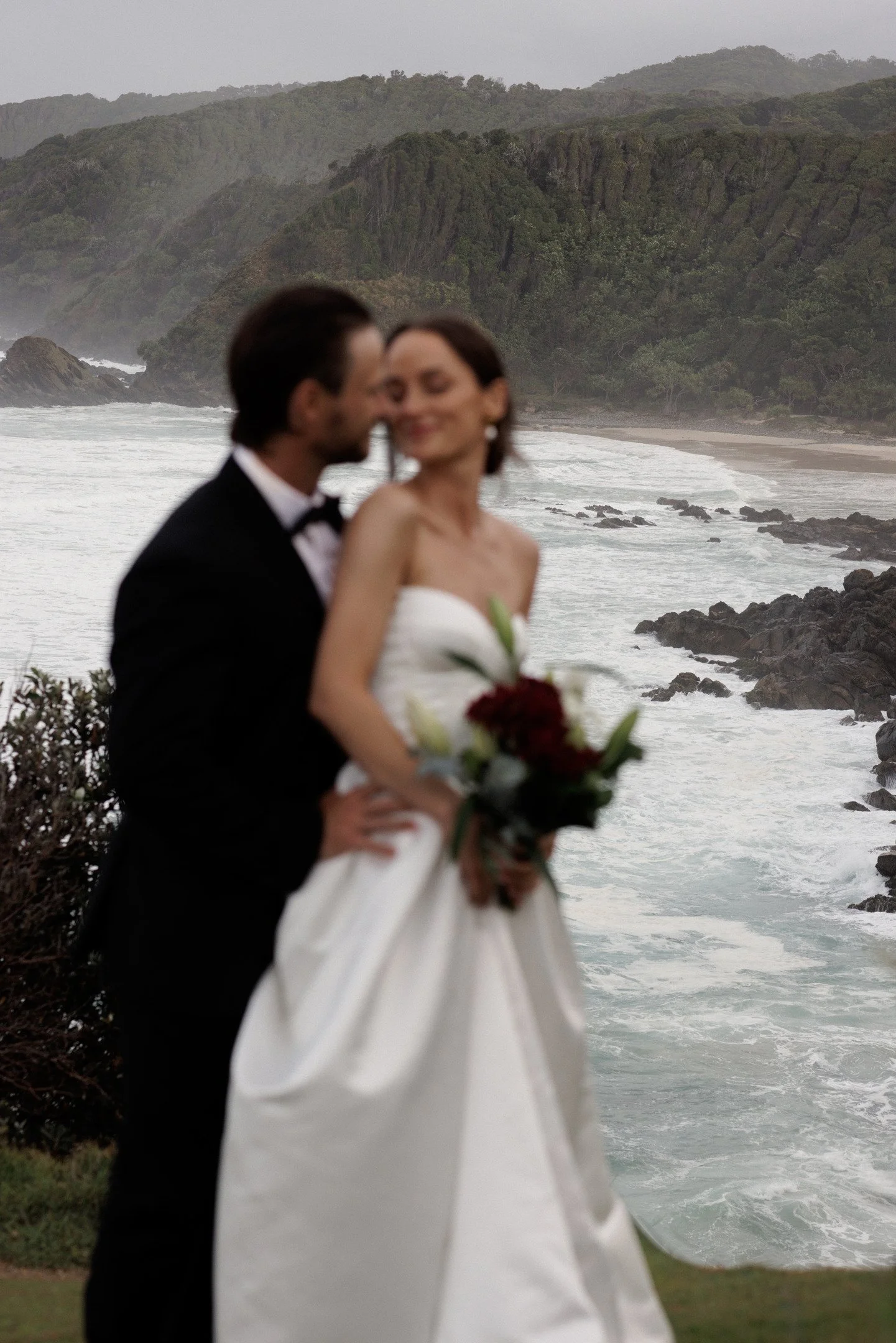A couple in wedding attire standing near the coast with waves crashing and green hills in the background.